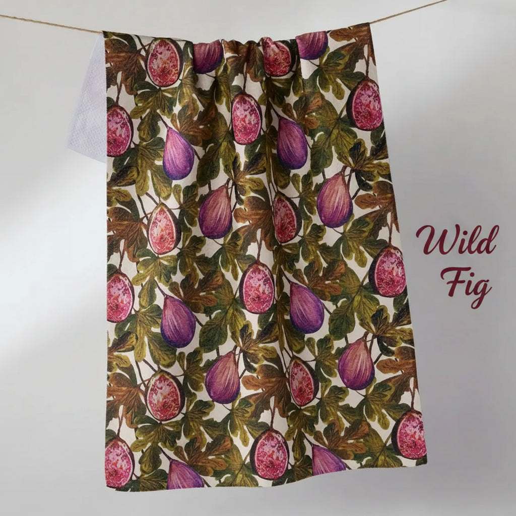 A Geometry wild fig tea towel with an all-over botanical print of purple figs and green leaves on a cream background, hanging from a thin cord against a light gray wall.