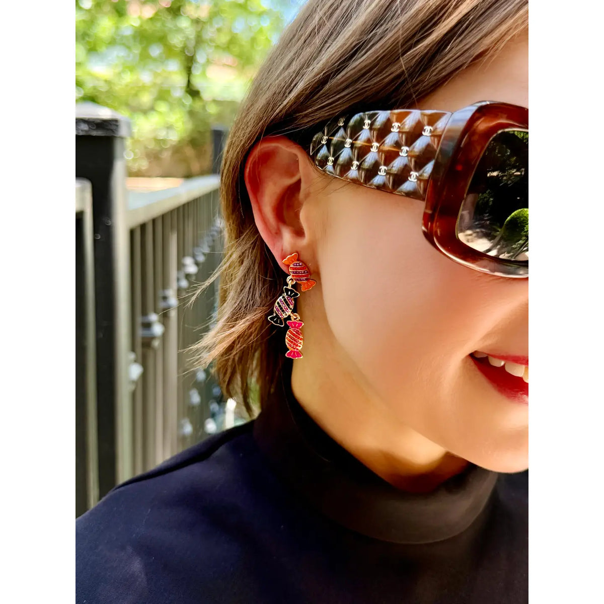 Woman with sunglasses wearing Canvas style drop earrings with 3 pieces of wrapped candy in orange, black and pink with purple, orange, pink and black pave stones.