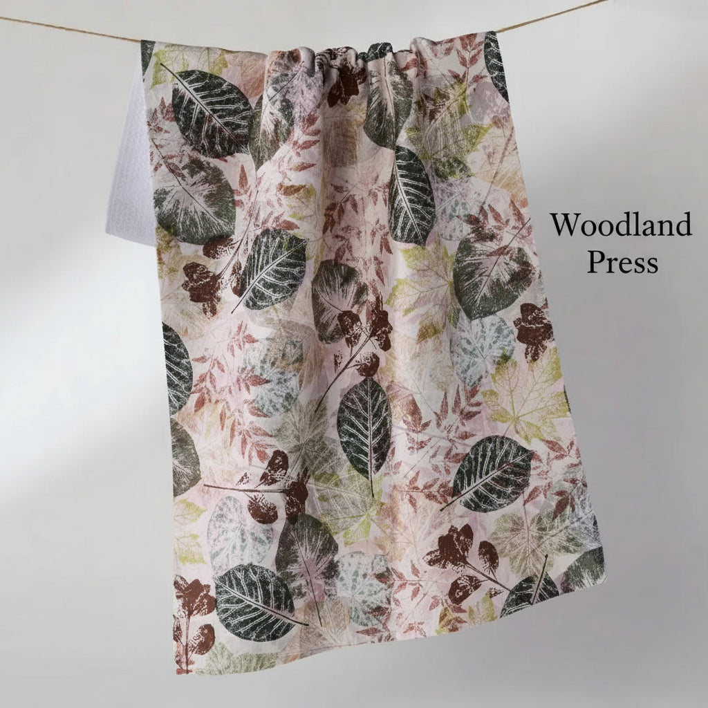 A fabric Geometry woodland press tea towel with a botanical leaf pattern in muted green, brown, and dusty pink, hanging from a thin cord against a light gray background.