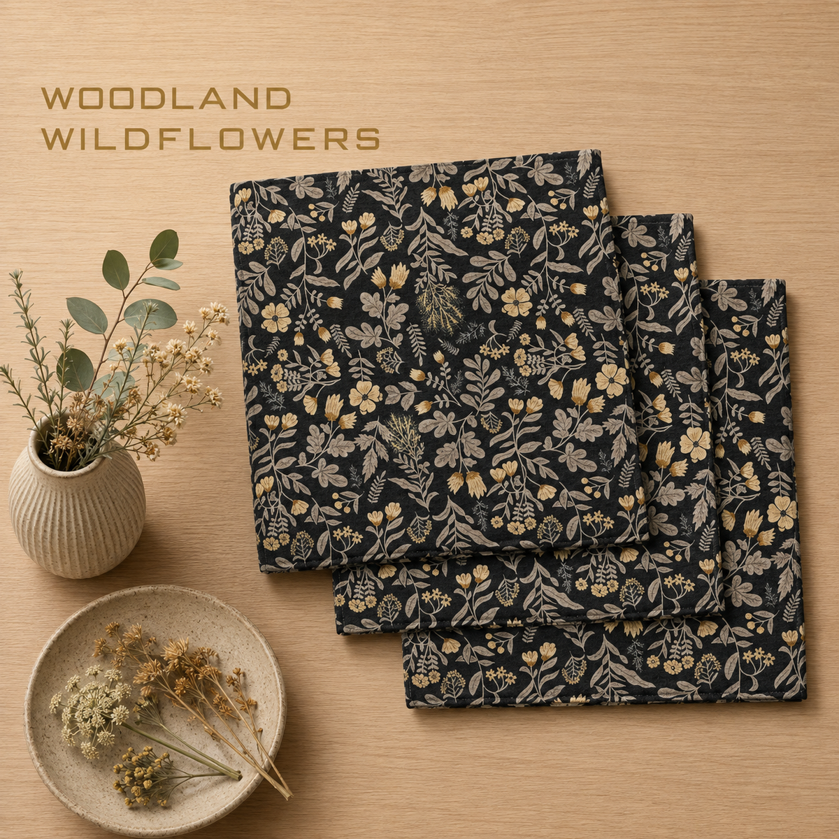 Geometry Woodland wildflowers dishcloth set with three square fabric dishcloths with a woodland floral pattern in black, beige, and muted yellow, arranged on a light wood tabletop next to a small vase of dried flowers and a plate of dried botanicals. The text 'WOODLAND WILDFLOWERS' appears in the upper left.  The Little Exchange SKU 142510