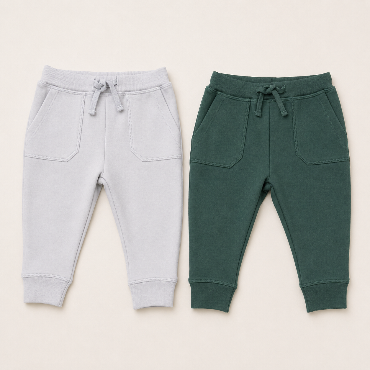 Two pairs of children's Angel Dear jogger sweatpants laid flat on a light beige background. The pair on the left is light gray, and the pair on the right is dark green.