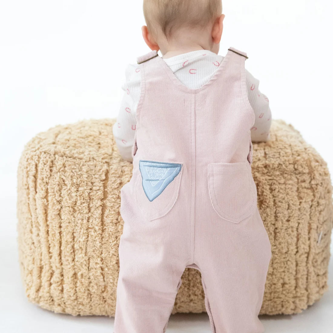 A baby seen from behind wearing light pink corduroy overalls  with a blue bandana applique on one pocket over a white long-sleeve shirt.