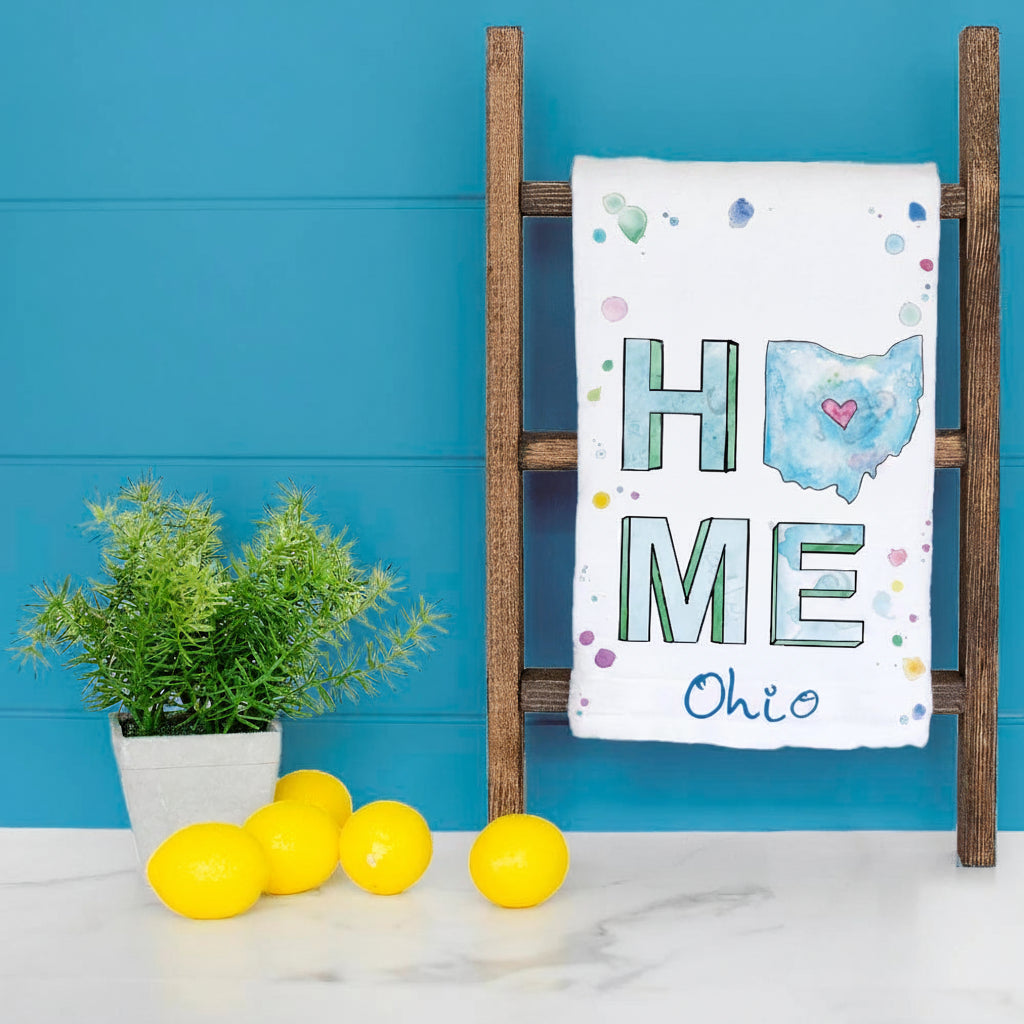 A white decorative towel draped over a rustic wooden ladder, featuring the word 'HOME' with 'O' as a blue map of Ohio and 'Ohio' written below.