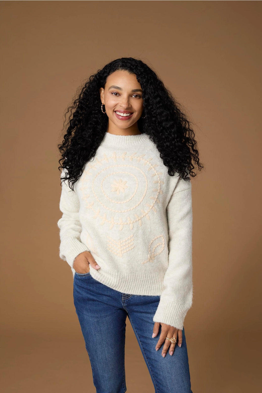 A woman wearing a cream-colored knit sweater with a light peach circular graphic on the front, paired with blue jeans, against a solid brown studio background.