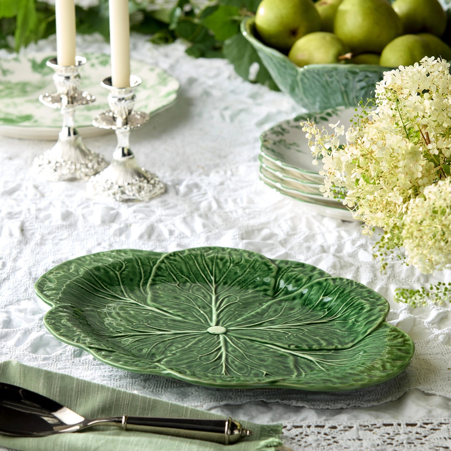 Green leaf-shaped ceramic plate on a table with white lace tablecloth, green apples, and white flowers.