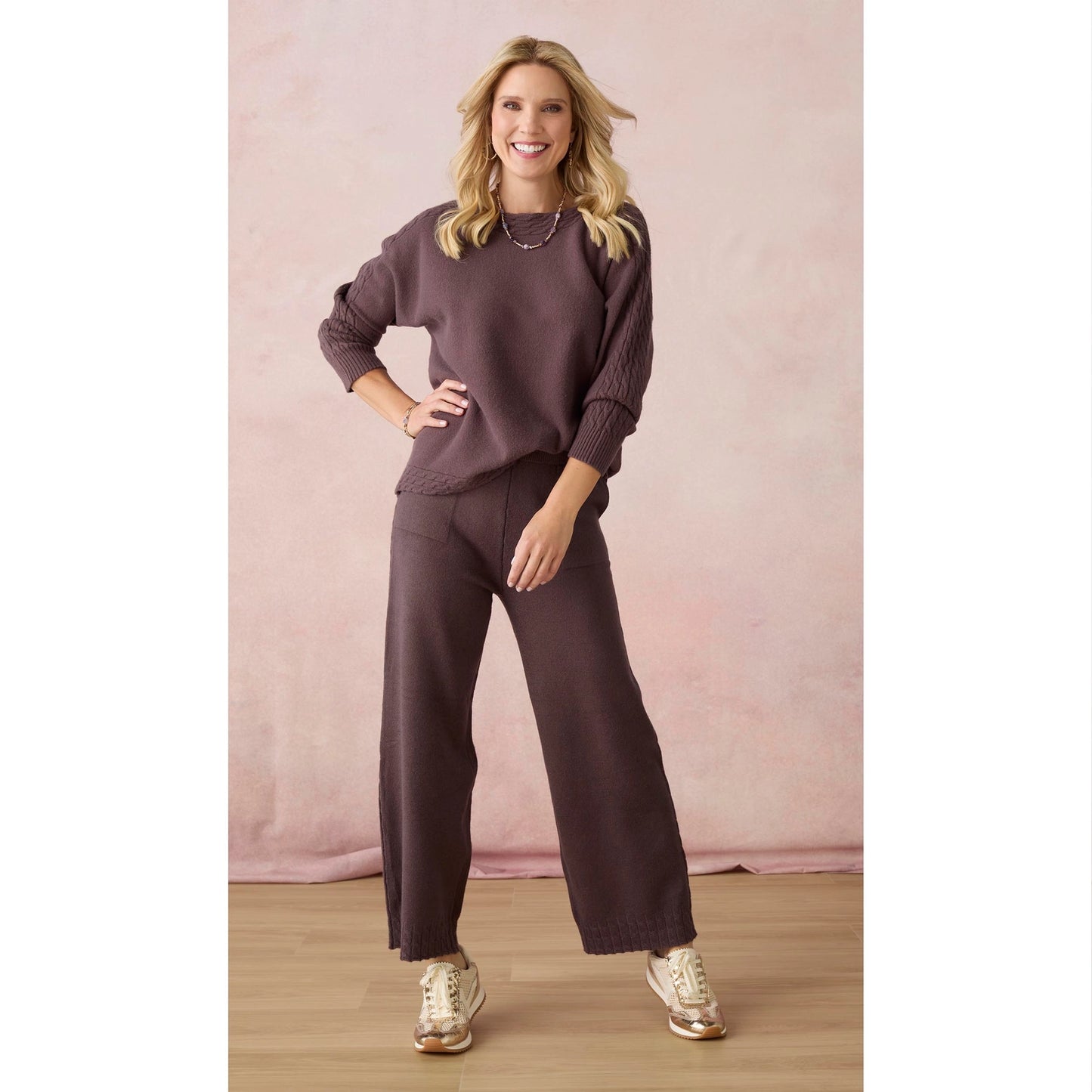 A woman modeling a matching two-piece lounge set consisting of a long-sleeve crewneck sweatshirt and wide-leg pants in a dusty plum purple color, paired with gold sneakers against a soft pink backdrop.