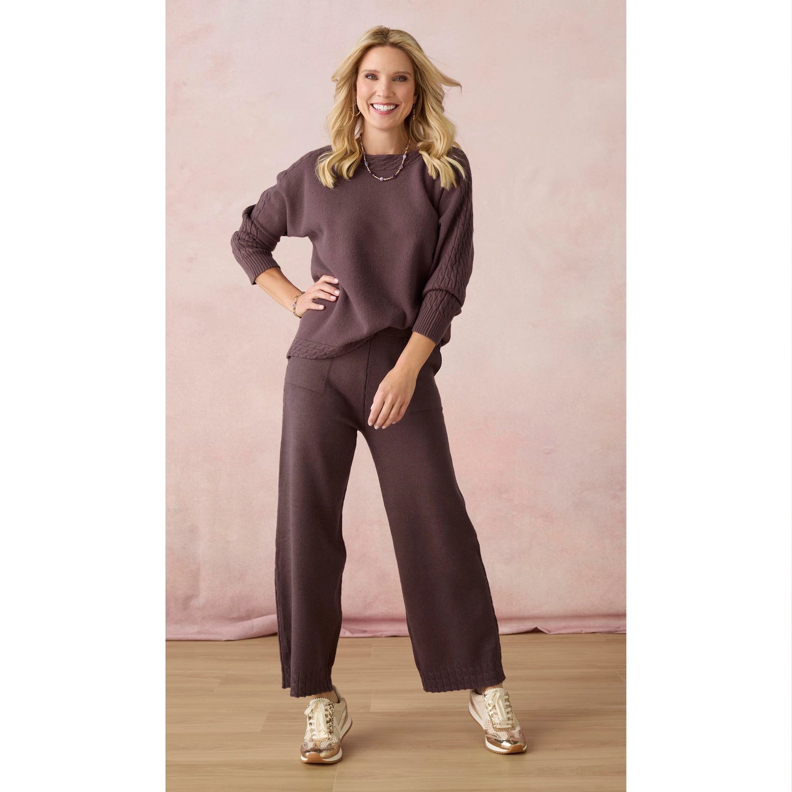 A woman modeling a matching two-piece lounge set consisting of a long-sleeve crewneck sweatshirt and wide-leg pants in a dusty plum purple color, paired with gold sneakers against a soft pink backdrop.