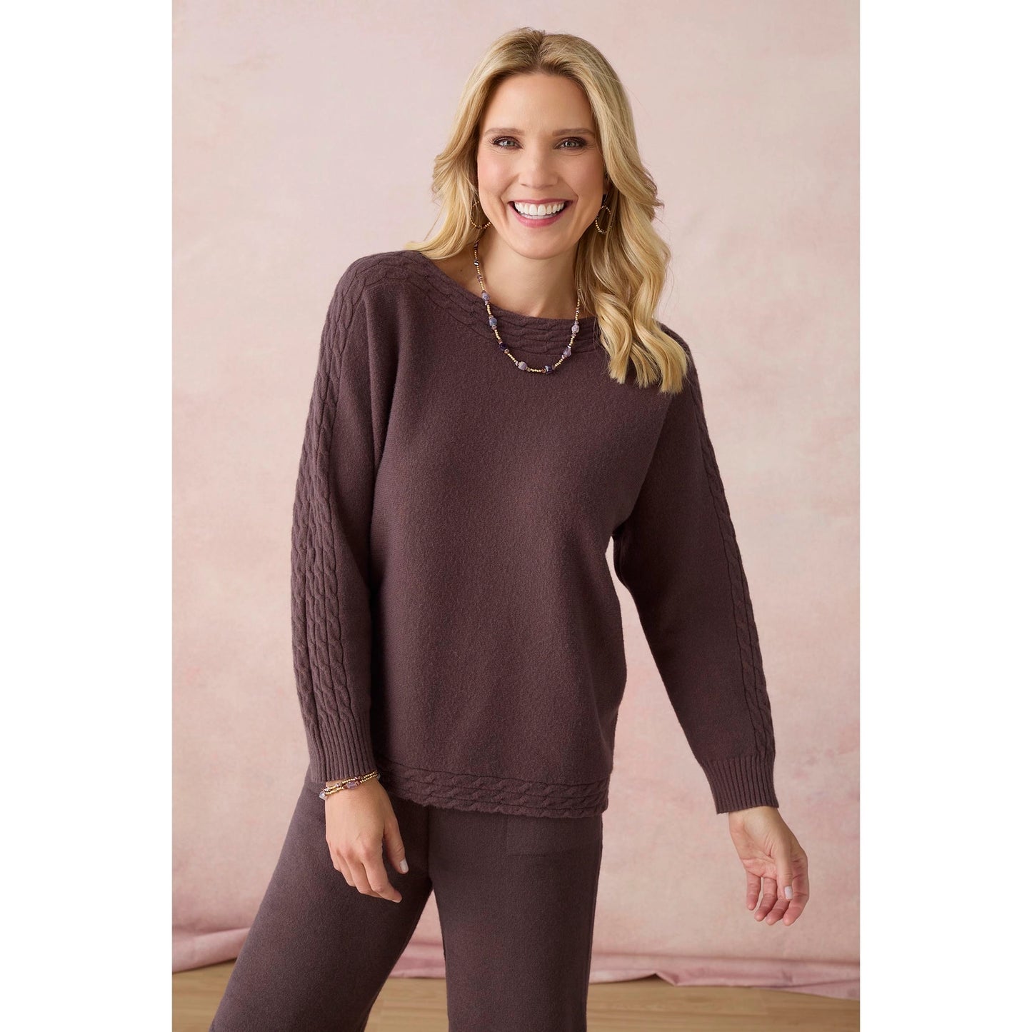 A smiling blonde woman models a dark brown cable-knit sweater and matching knit pants against a soft pink studio backdrop.