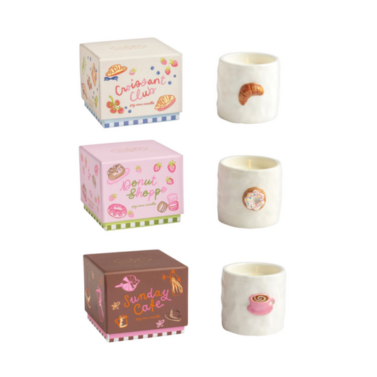 Three small ceramic candle holders with matching decorative boxes, each featuring a different bakery-themed design: Crispy Claws, Donut Shop, and Sunday Cake. The candles are off-white with colorful illustrations of pastries and coffee, shown on a plain white background.