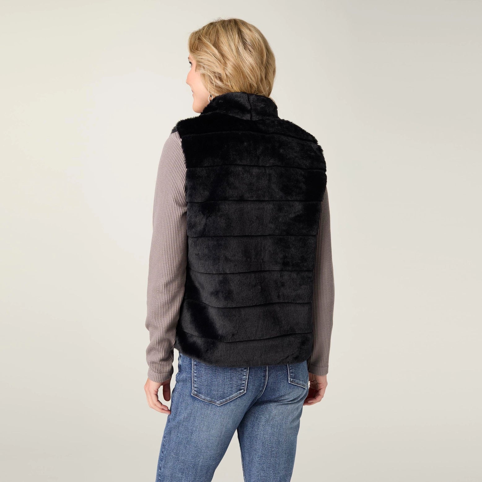 A woman shown from the back wearing a black faux fur vest over a ribbed long-sleeve top and blue jeans, photographed against a plain light gray studio background.