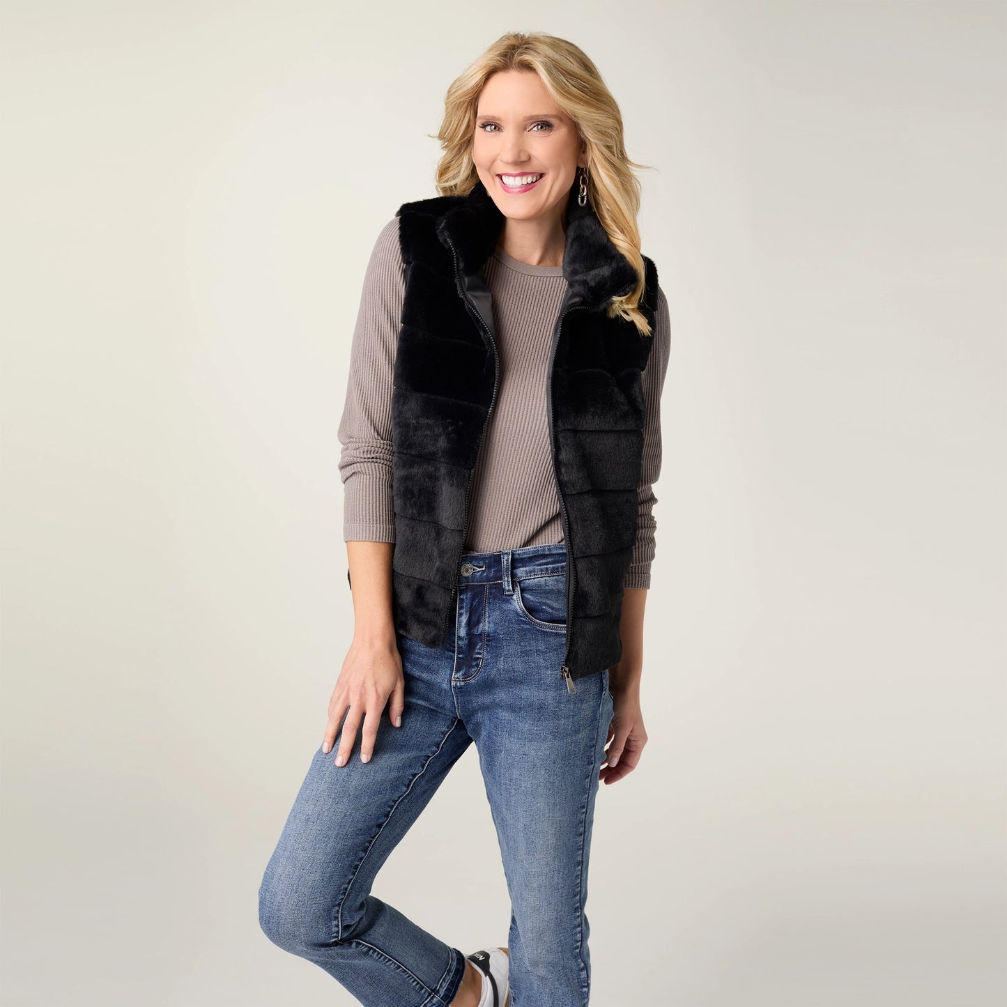 A woman modeling a black faux-fur vest over a taupe ribbed long-sleeve top and blue jeans, standing against a plain light gray studio background.