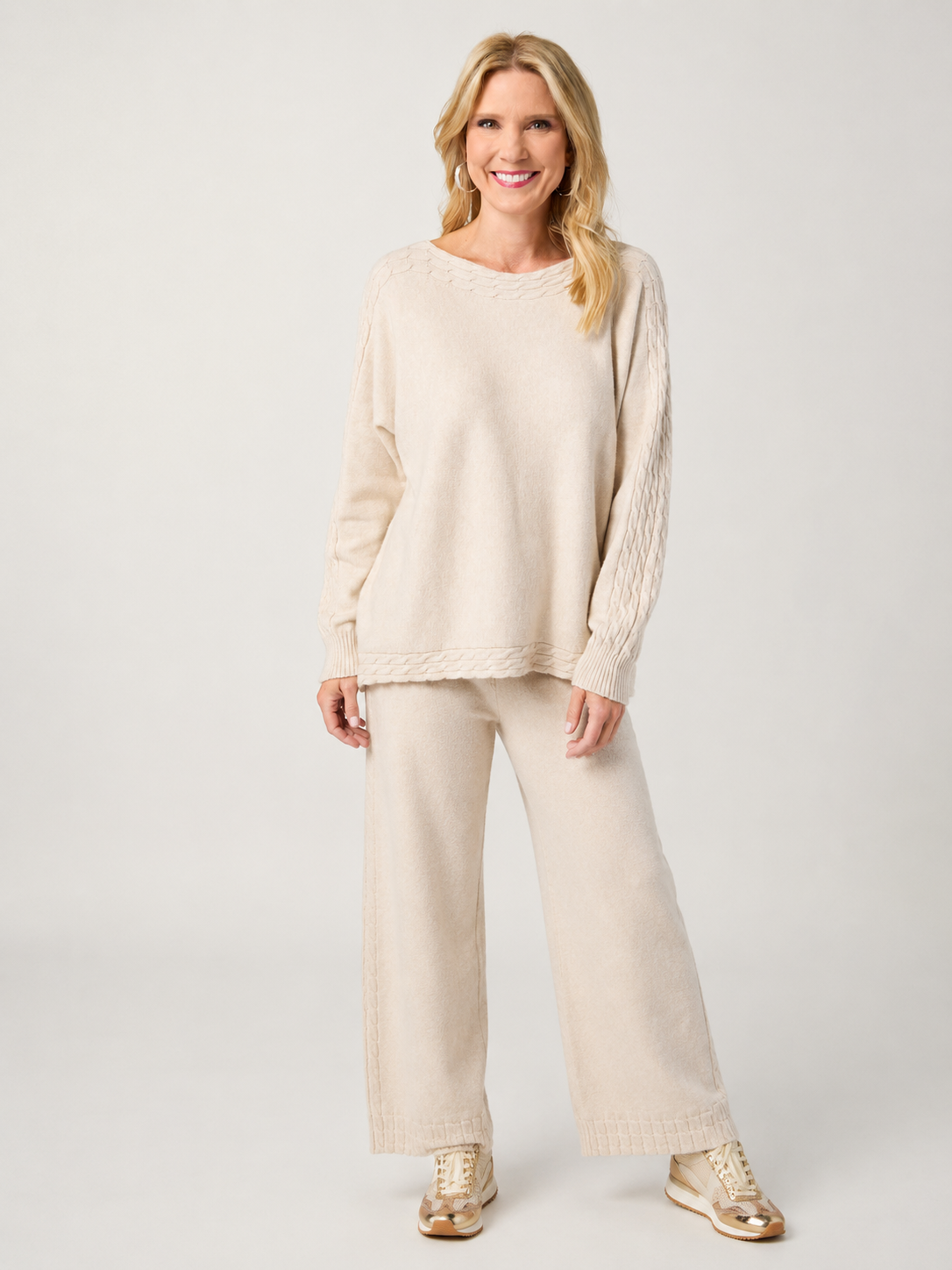 A woman modeling a matching knit two-piece set consisting of a cream-colored long-sleeve top and wide-leg pants, paired with metallic gold sneakers, against a light gray studio background.