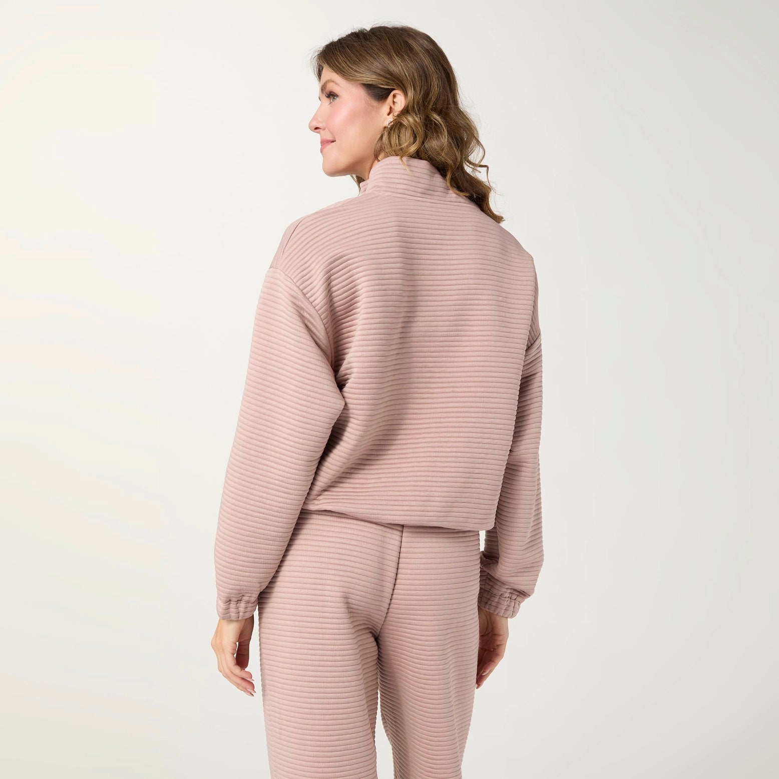 A woman shown from the back wearing a matching ribbed knit lounge set in a dusty pink color, consisting of a long-sleeve top and pants, against a plain light background.
