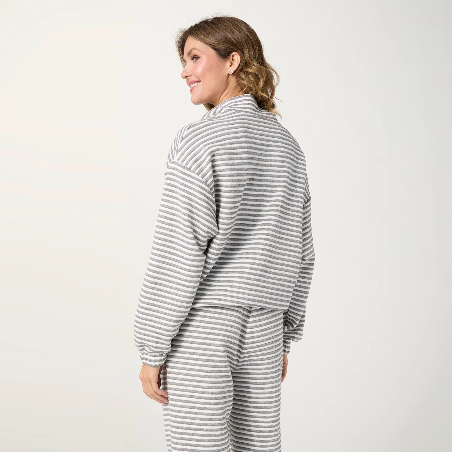 A woman wearing a matching gray and white striped lounge set, shown from a side-back angle against a plain light gray studio background.