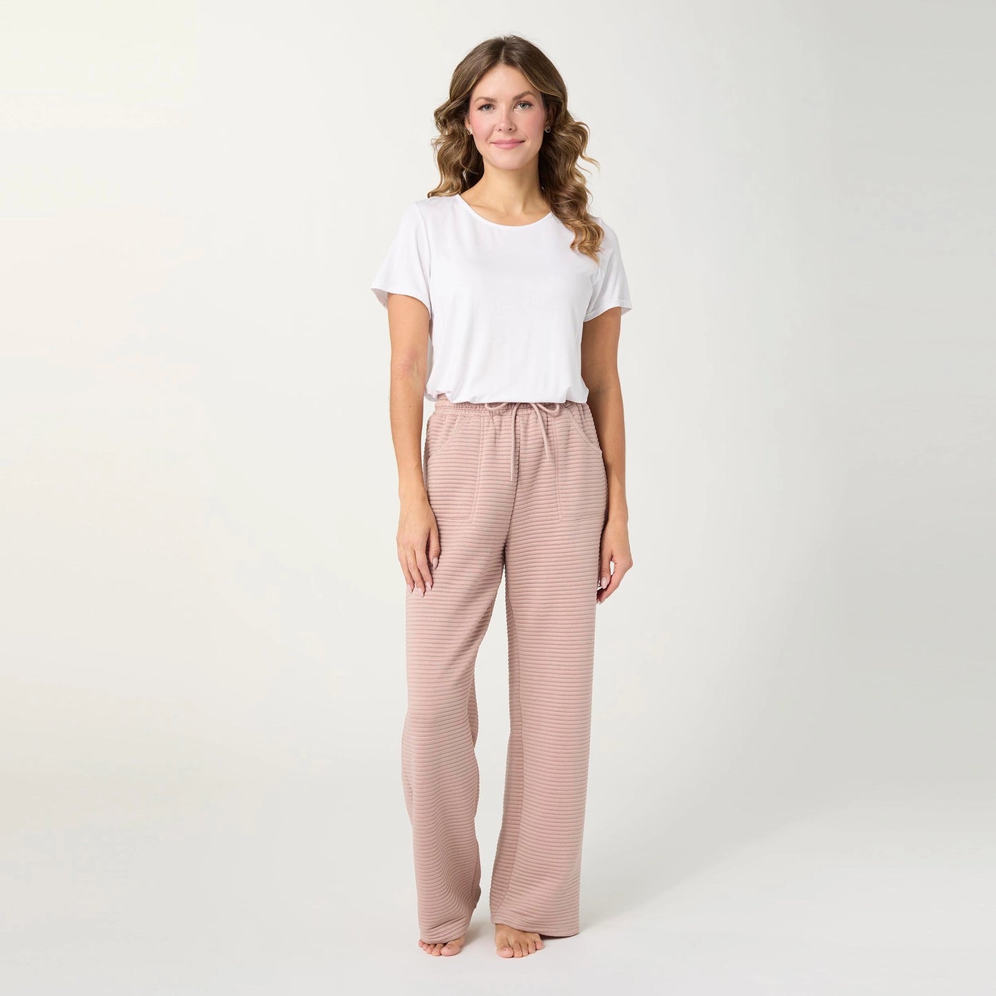 A woman stands barefoot against a plain light gray studio background, wearing a white short-sleeve T-shirt and loose, wide-leg drawstring pants in a muted dusty pink or mauve tone.