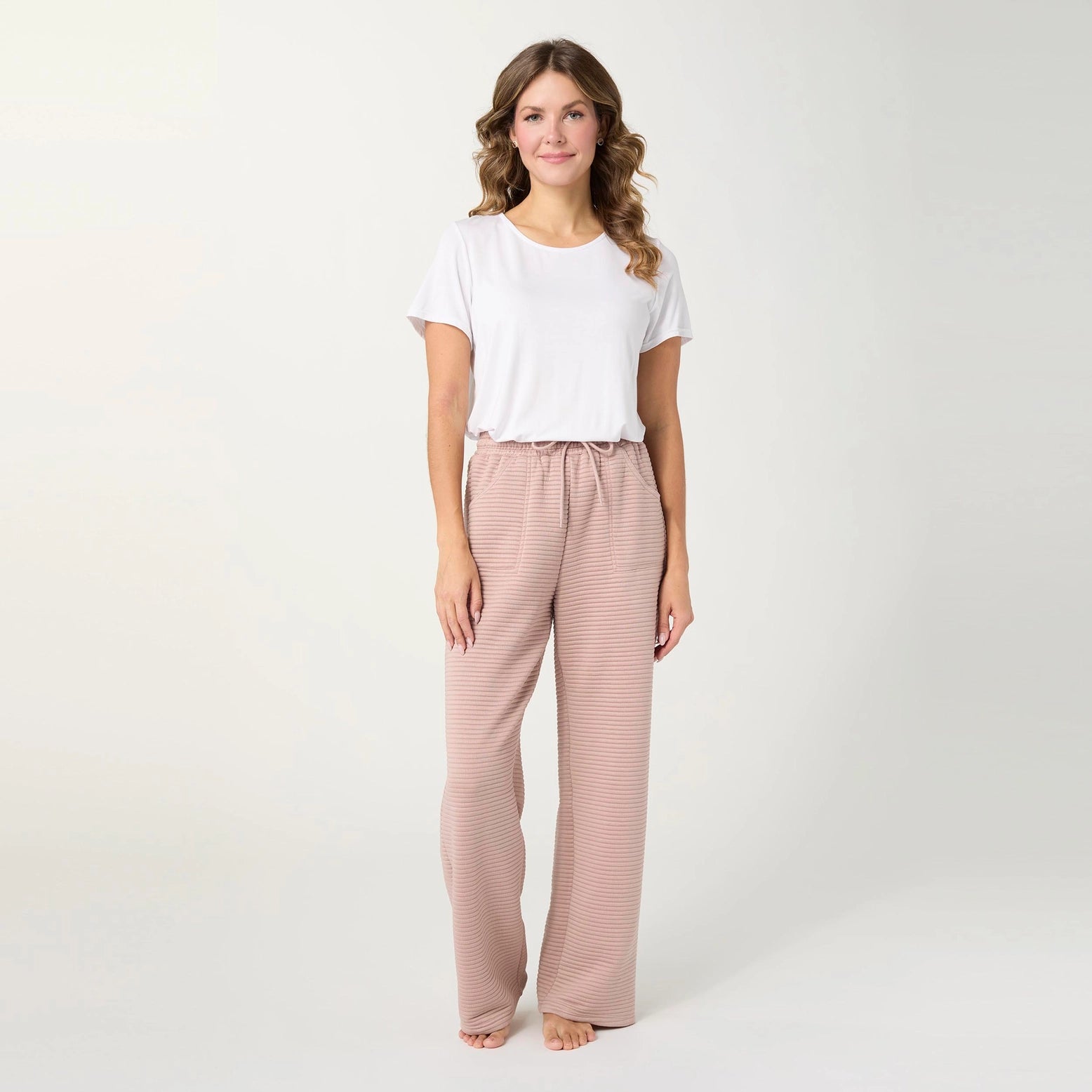 A woman stands barefoot against a plain light gray studio background, wearing a white short-sleeve T-shirt and loose, wide-leg drawstring pants in a muted dusty pink or mauve tone.