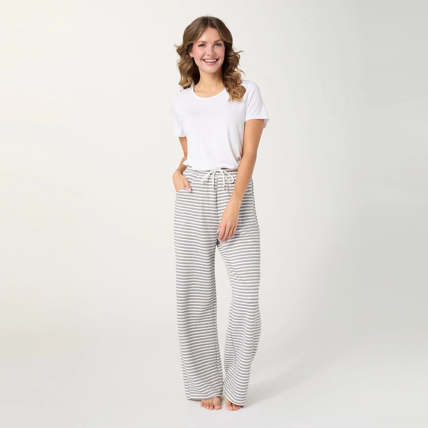 A woman stands barefoot against a light gray studio background, wearing a white short-sleeve t-shirt and white lounge pants with thin gray horizontal stripes and a drawstring waist.