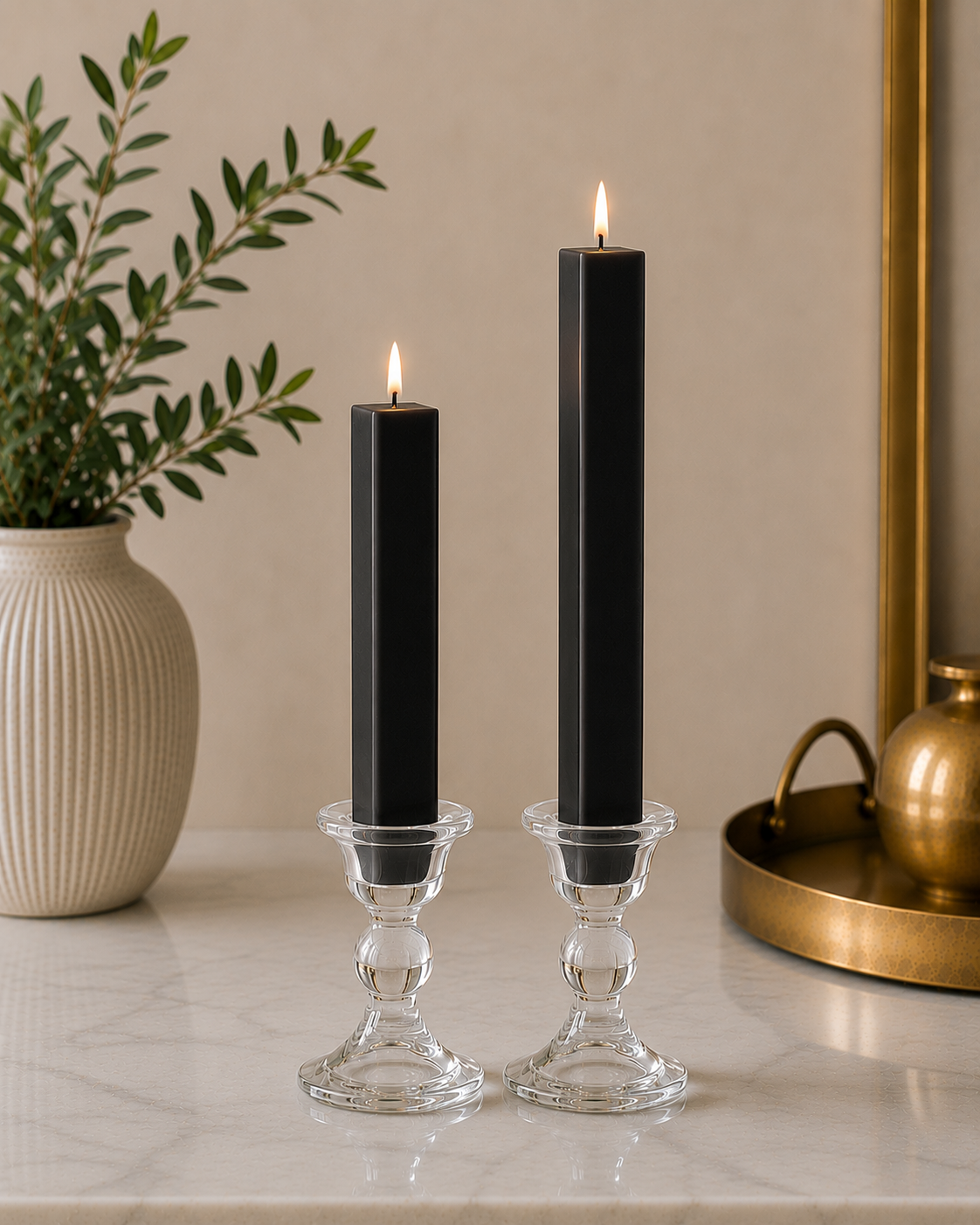 Two #34 Black 8" and 12" square taper pillar beeswax candles by Dadant  in clear glass candlesticks on a marble surface, with a potted plant on the left and a brass tray on the right.