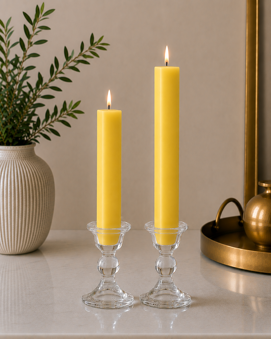 Two #74 Citrus Yellow 8" and 12" square taper pillar beeswax candles by Dadant  in clear glass candlesticks on a marble surface, with a potted plant on the left and a brass tray on the right.