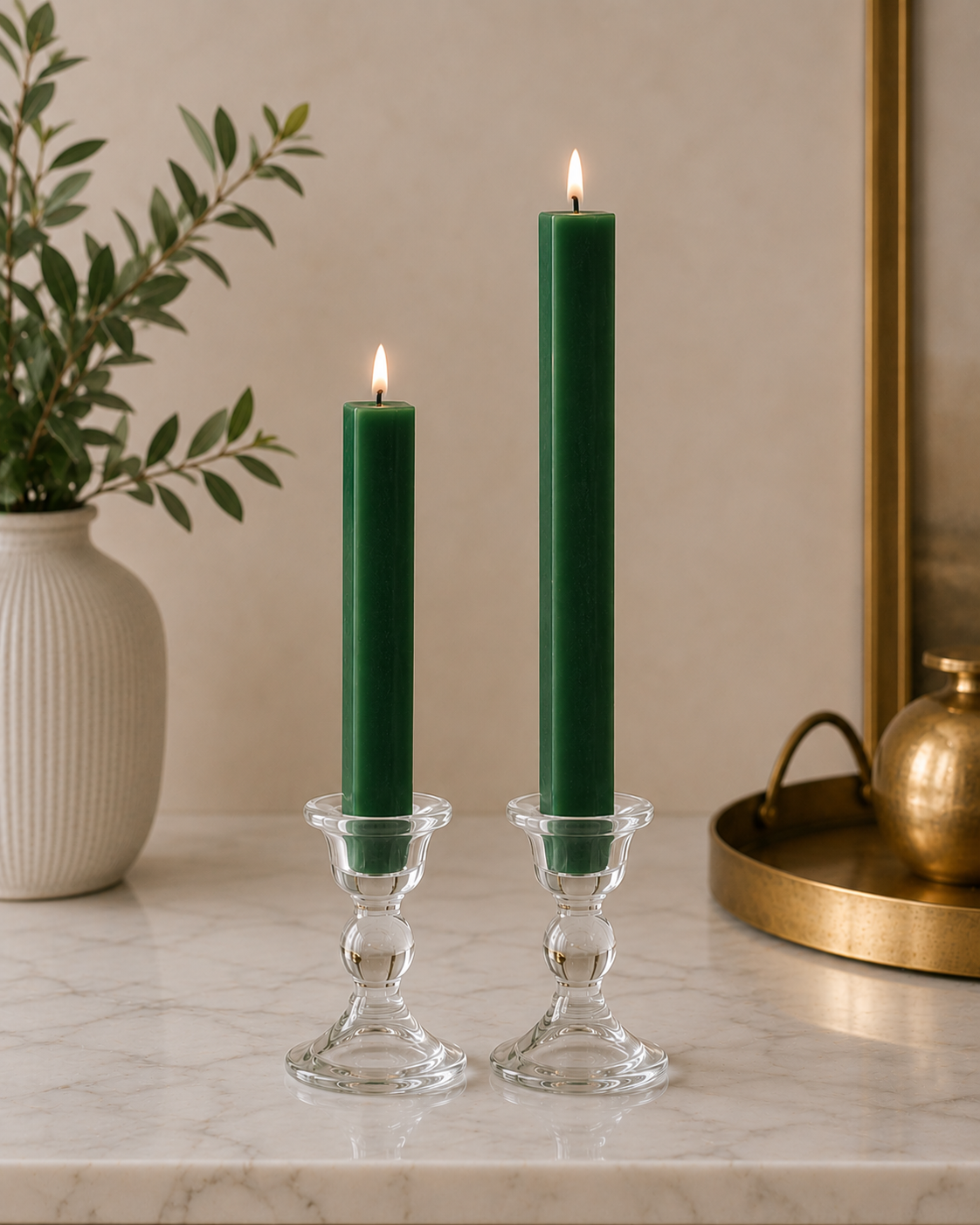 Two #14 forest green 8" and 12" square taper pillar beeswax candles by Dadant  in clear glass candlesticks on a marble surface, with a potted plant on the left and a brass tray on the right.