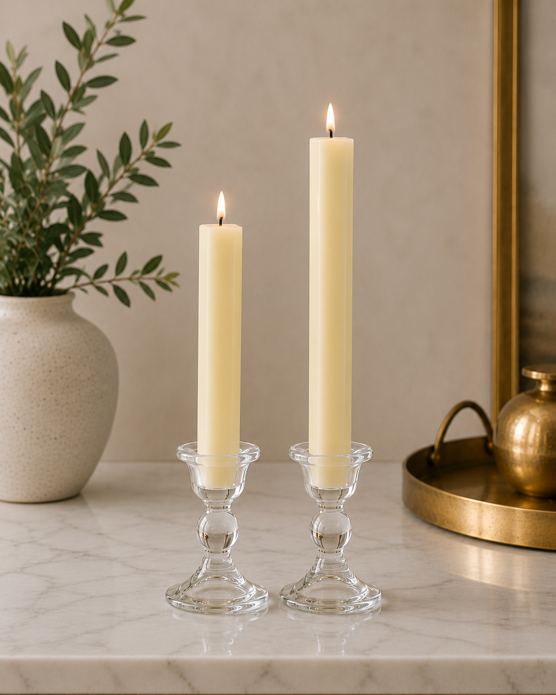 Two #43 french vanilla 8" and 12" square taper pillar beeswax candles by Dadant  in clear glass candlesticks on a marble surface, with a potted plant on the left and a brass tray on the right.