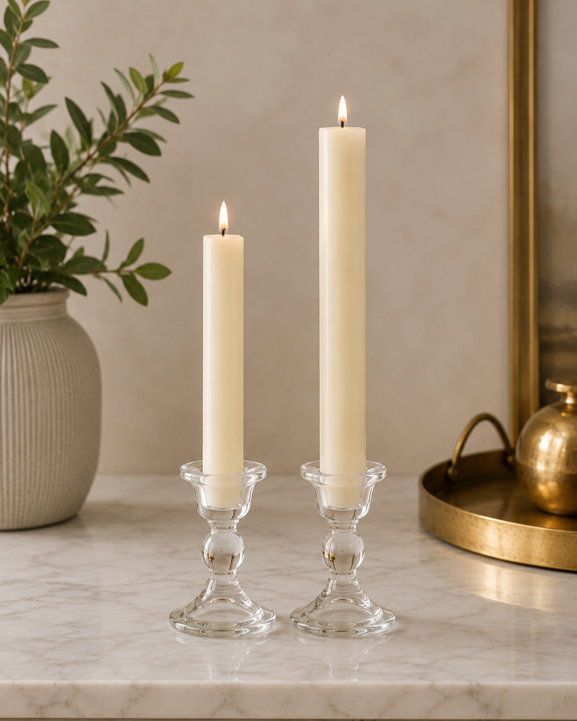 Two #03 linen white 8" and 12" square taper pillar beeswax candles by Dadant  in clear glass candlesticks on a marble surface, with a potted plant on the left and a brass tray on the right.