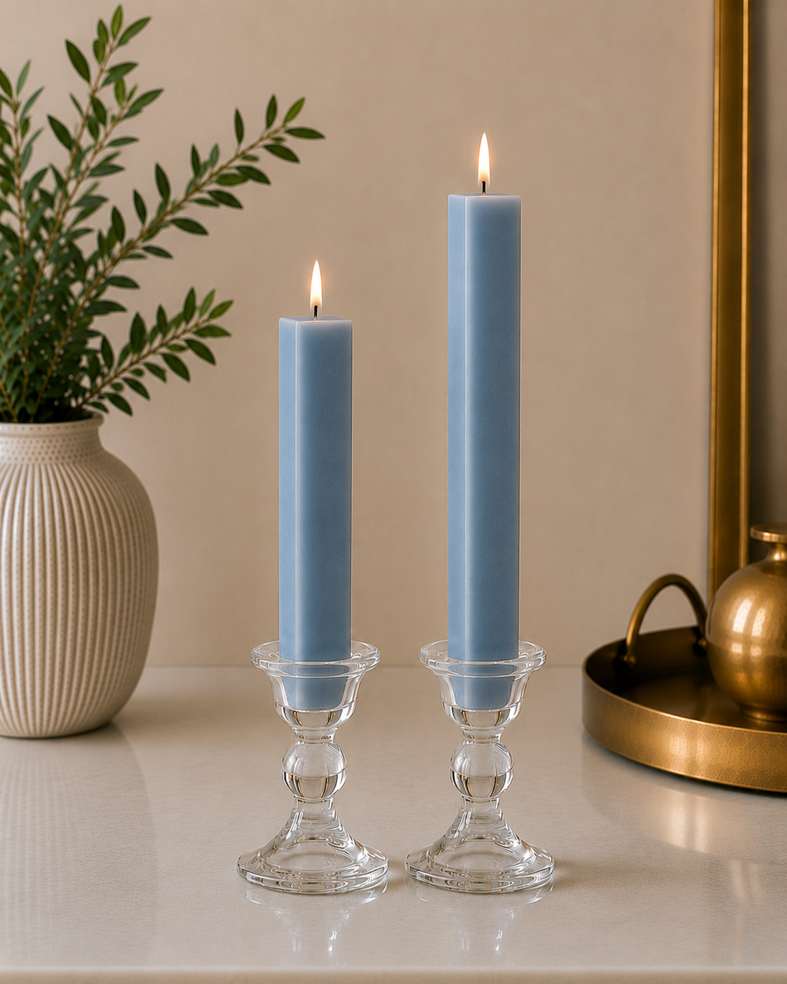 Two lit blue taper candles in clear glass candle holders on a tabletop, with a beige wall background, a potted green plant on the left, and a gold-toned decorative object on the right.