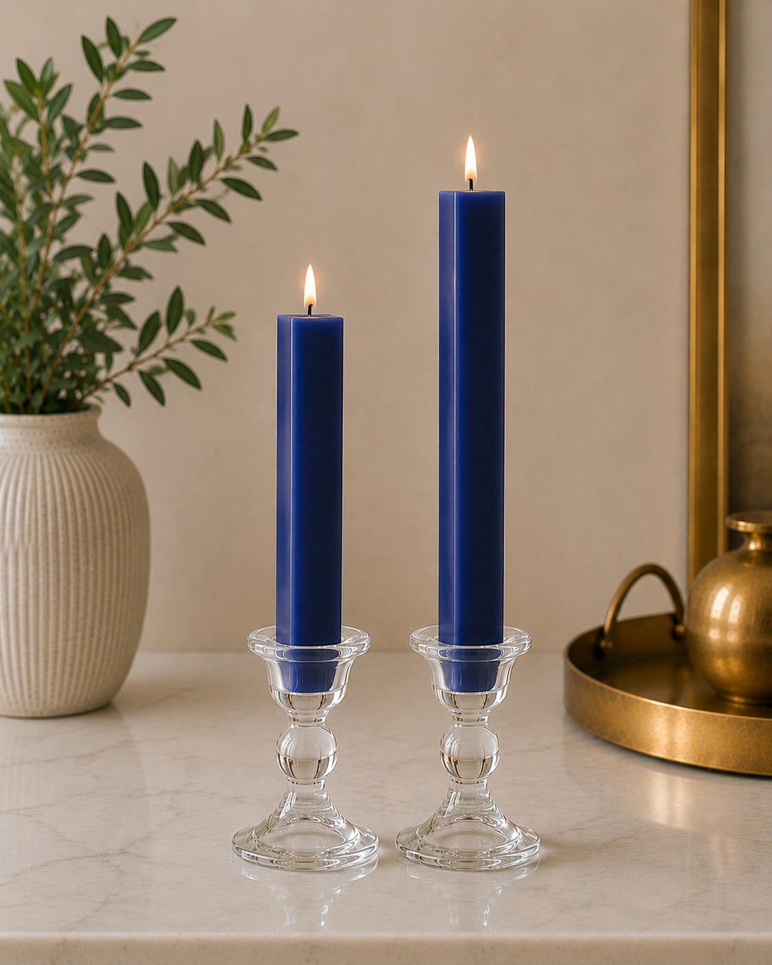 Two #19 Navy Blue 8" and 12" square taper pillar beeswax candles by Dadant  in clear glass candlesticks on a marble surface, with a potted plant on the left and a brass tray on the right.