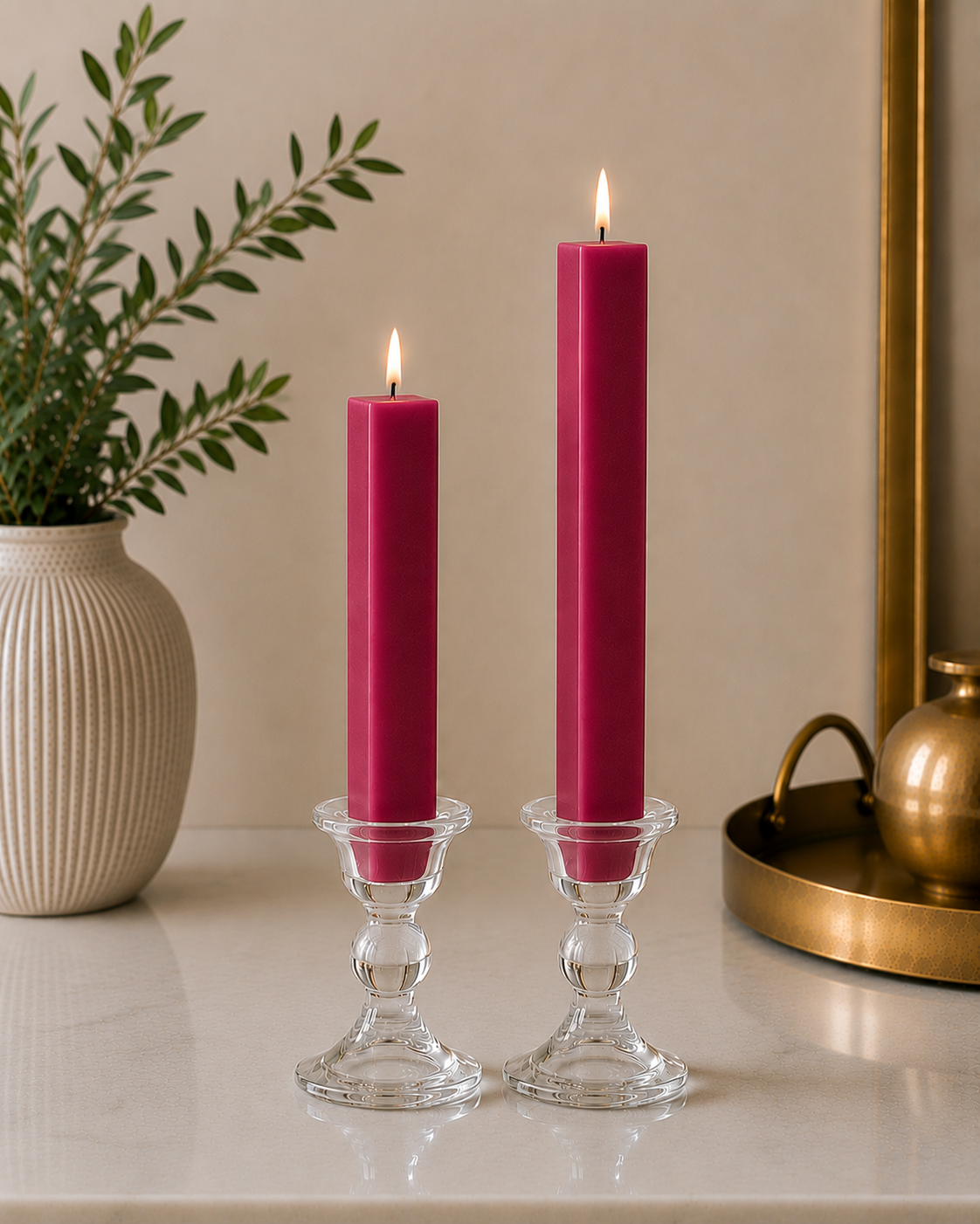 Two #69 Raspberry 8" and 12" square taper pillar beeswax candles by Dadant  in clear glass candlesticks on a marble surface, with a potted plant on the left and a brass tray on the right.