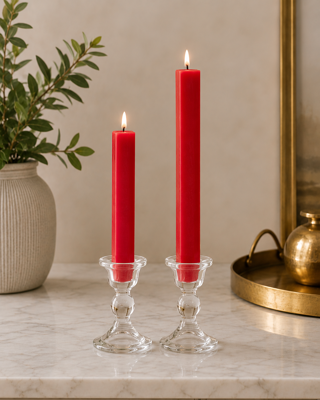 Two #12 real red 8" and 12" square taper pillar beeswax candles by Dadant  in clear glass candlesticks on a marble surface, with a potted plant on the left and a brass tray on the right.