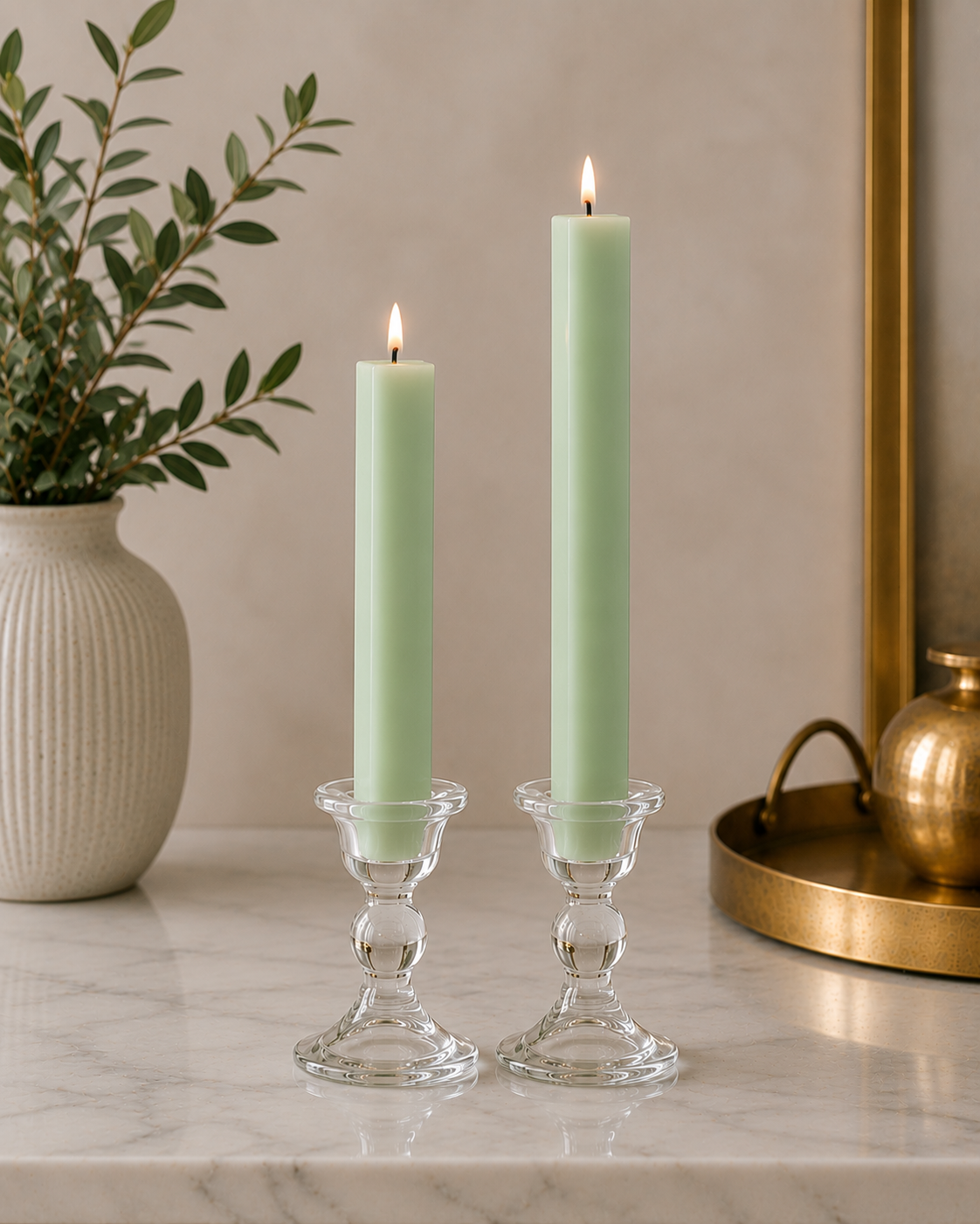 Two lit pale green taper candles in clear glass candlesticks on a marble surface, with a white vase of greenery on the left and a gold tray on the right.
