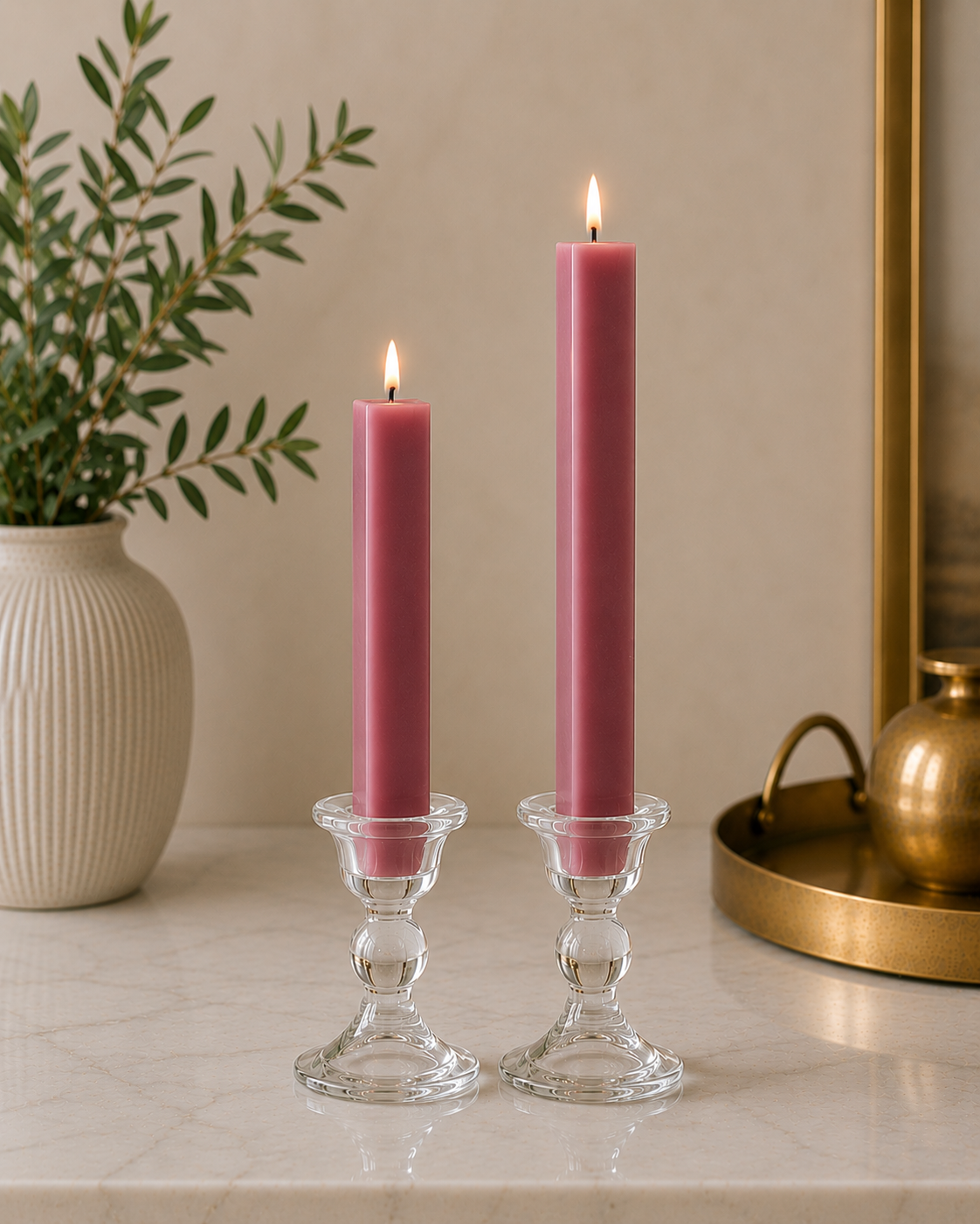 Two lit dadant beeswack square pink taper candles in clear glass candlesticks on a light marble surface, with a white ribbed vase and green leaves on the left and a gold tray on the right.