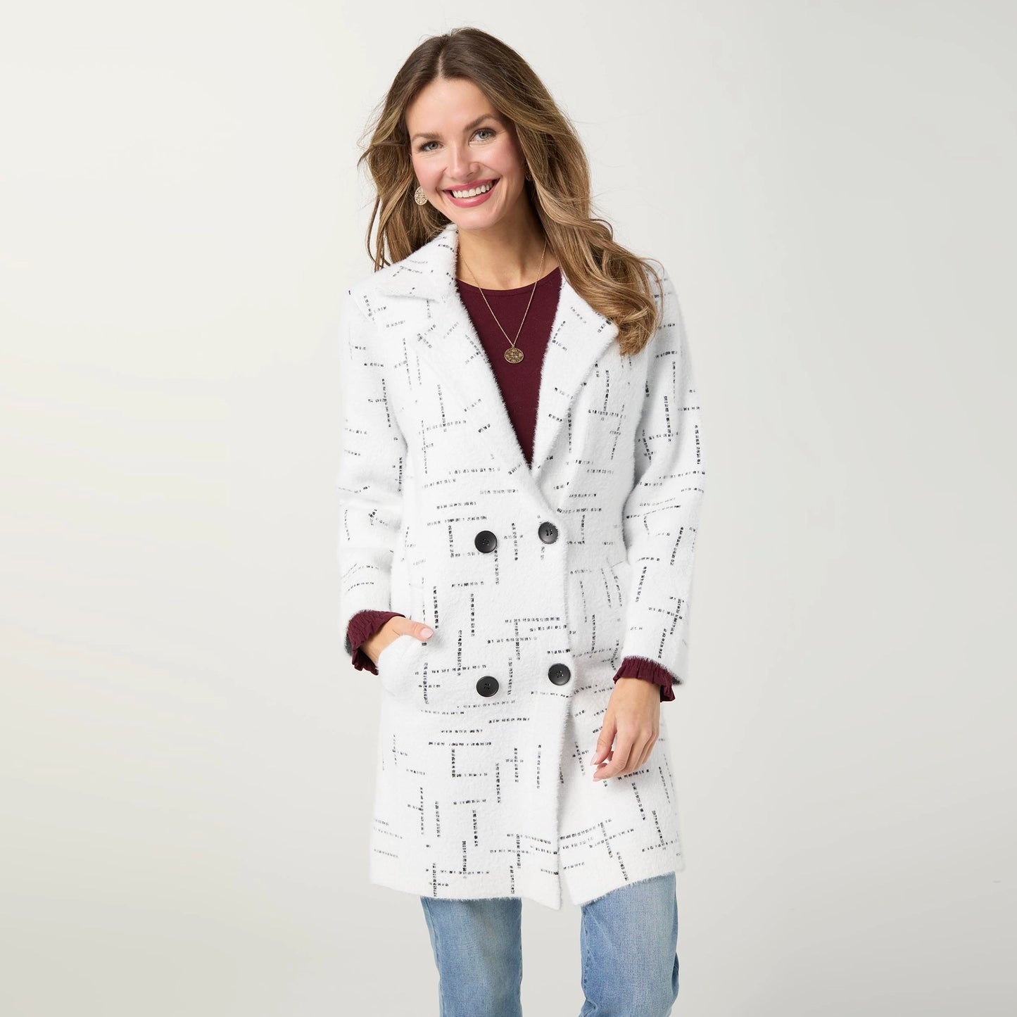 A woman modeling a white double-breasted cardigan with a black geometric line pattern, worn over a burgundy top and blue jeans against a light gray studio background.