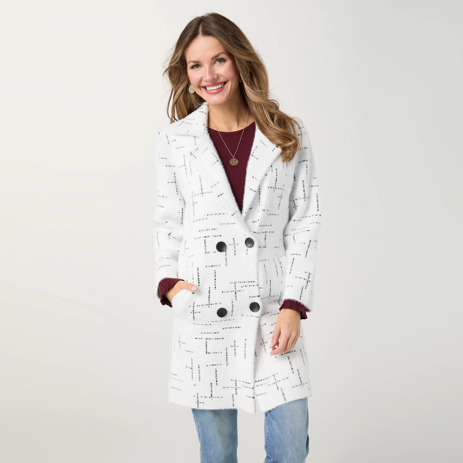A woman modeling a white double-breasted cardigan with a black geometric line pattern, worn over a burgundy top and blue jeans against a light gray studio background.