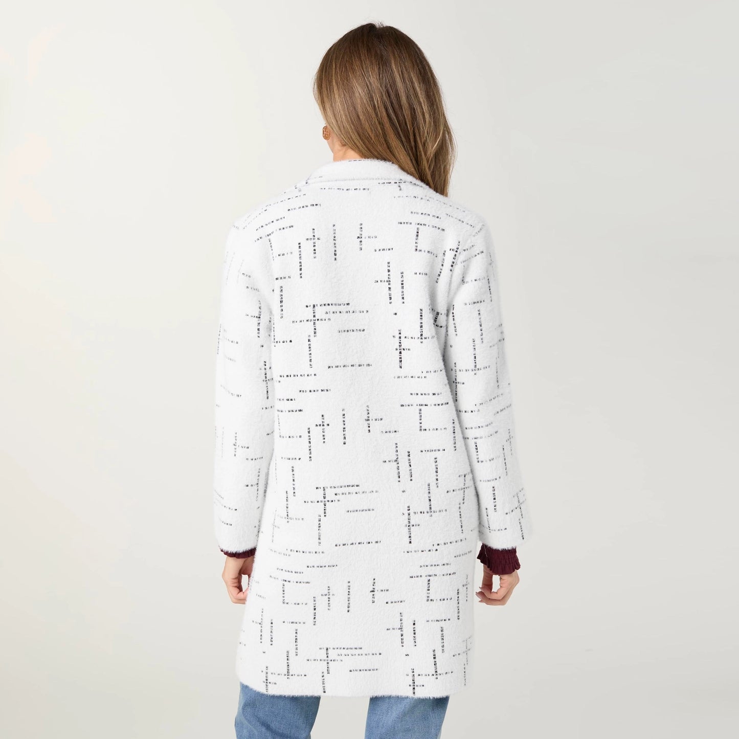 A woman viewed from behind wearing a white long coat with a black typographic pattern and dark burgundy cuffs, paired with blue jeans, against a plain light gray studio background.