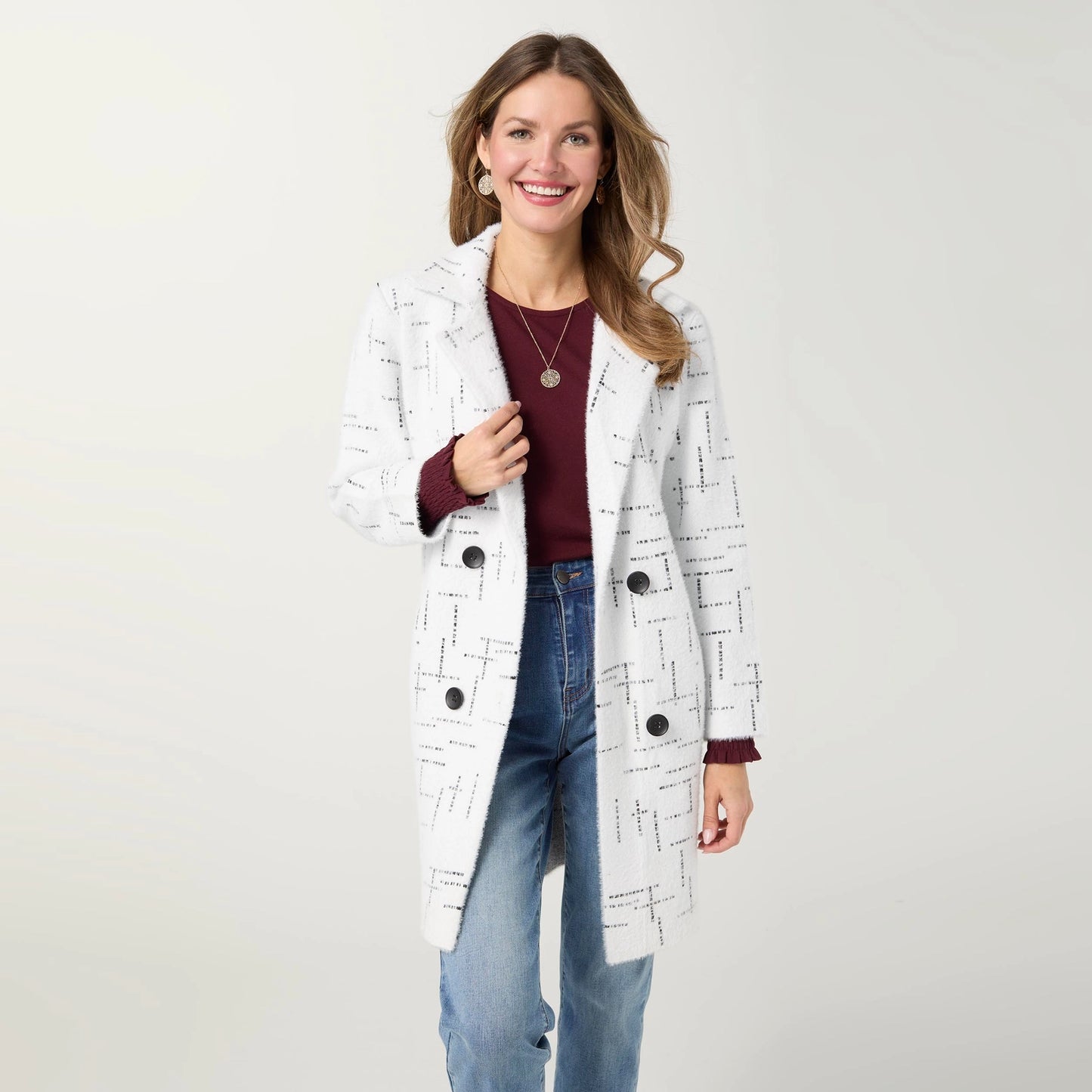 A woman modeling a white double-breasted cardigan with a black geometric line pattern, worn over a burgundy top and blue jeans against a light gray studio background.