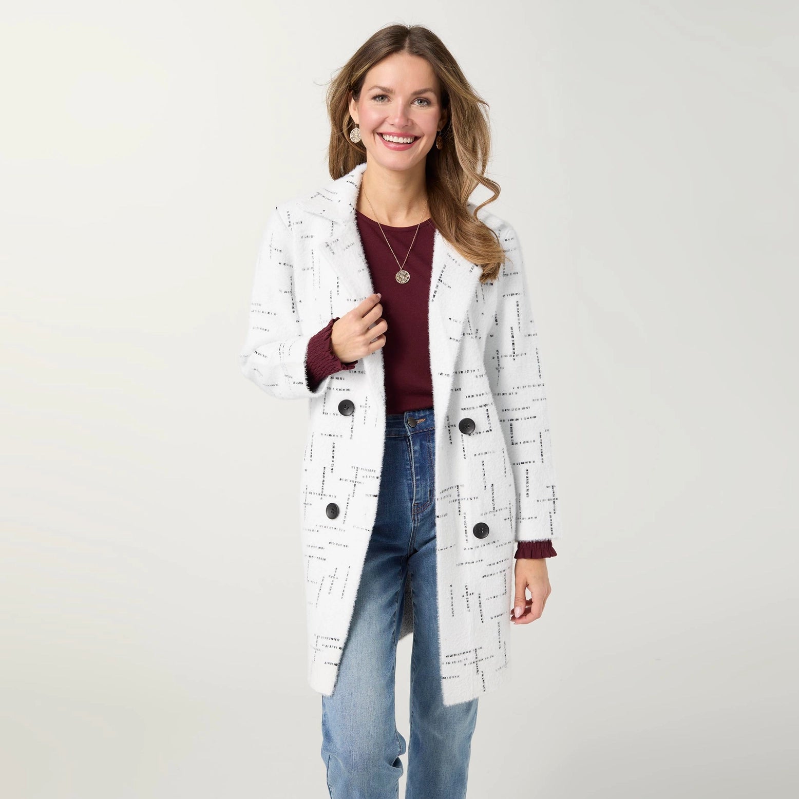 A woman modeling a white double-breasted cardigan with a black geometric line pattern, worn over a burgundy top and blue jeans against a light gray studio background.