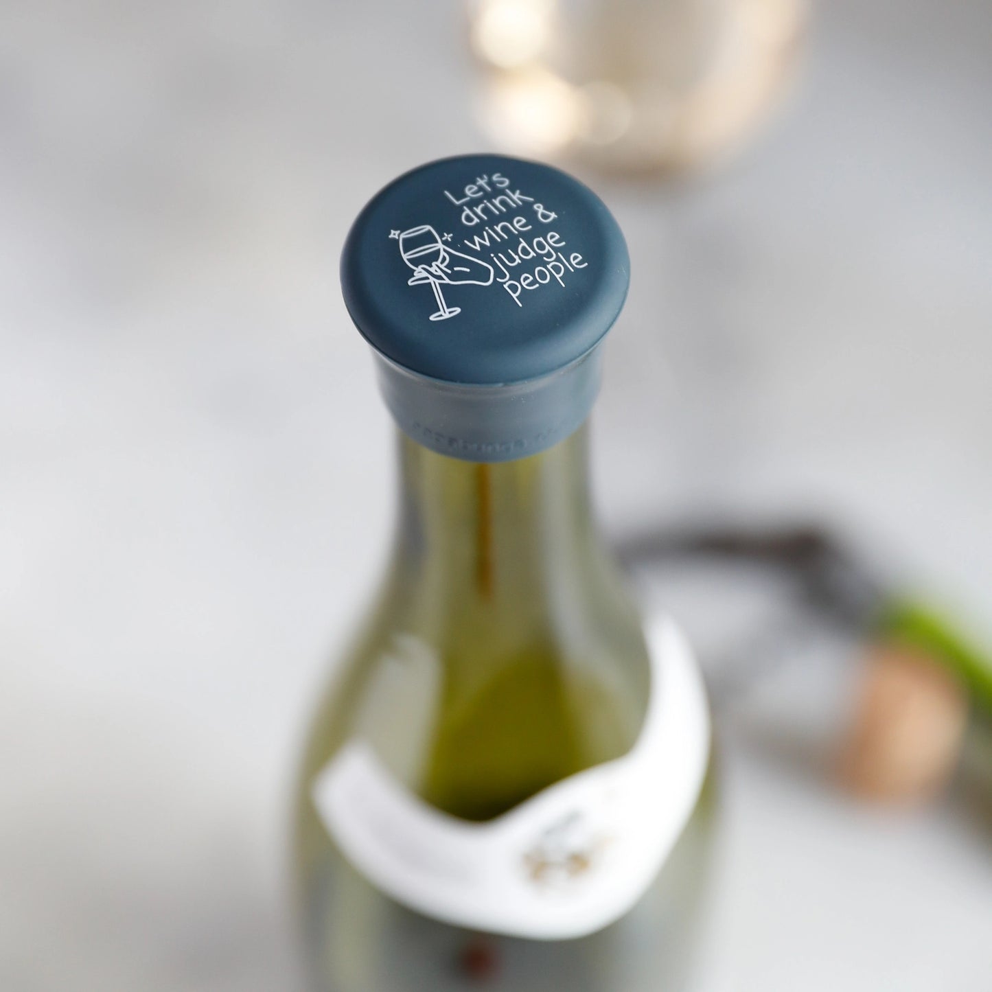 Wine bottle with a blue cork labeled 'Let's drink wine & judge people' on a blurred background.