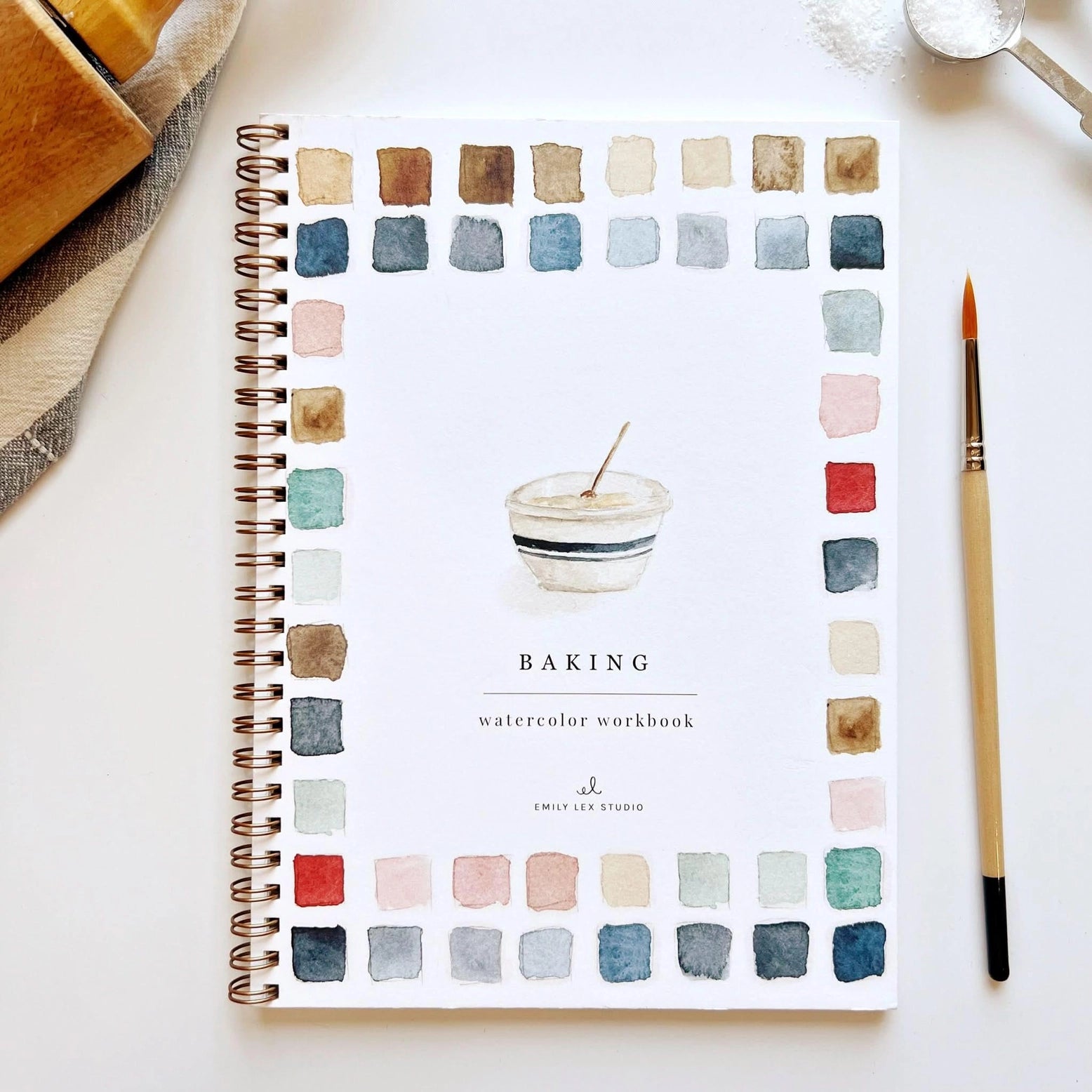 Baking watercolor workbook with color swatches and a mixing bowl on a white surface with art supplies.
