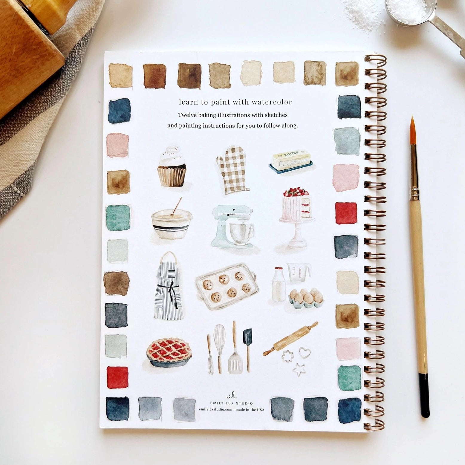Notebook with watercolor baking illustrations and a paintbrush on a white surface