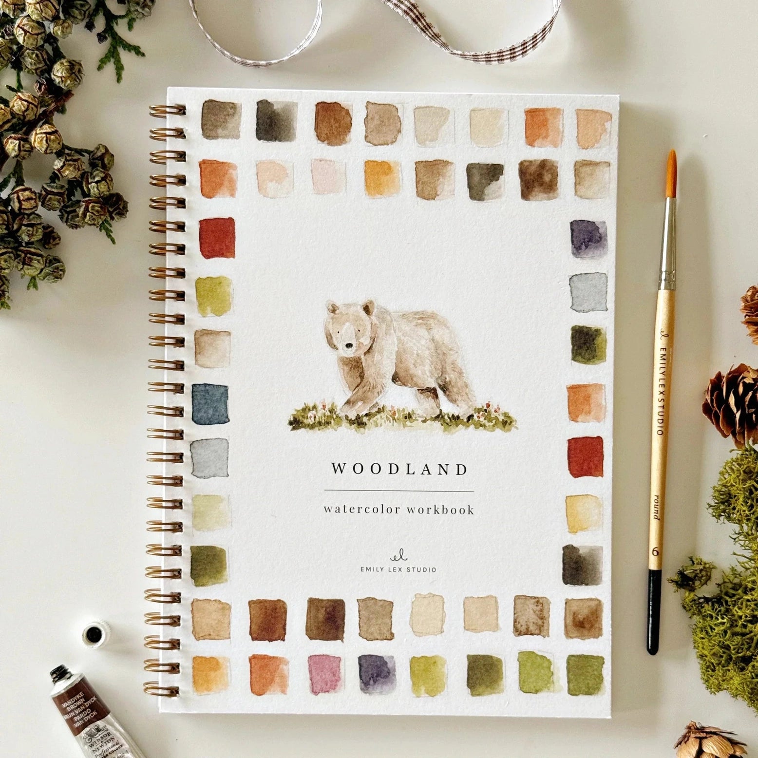 Woodland watercolor workbook with color swatches and a brown bear on a white surface with art supplies.