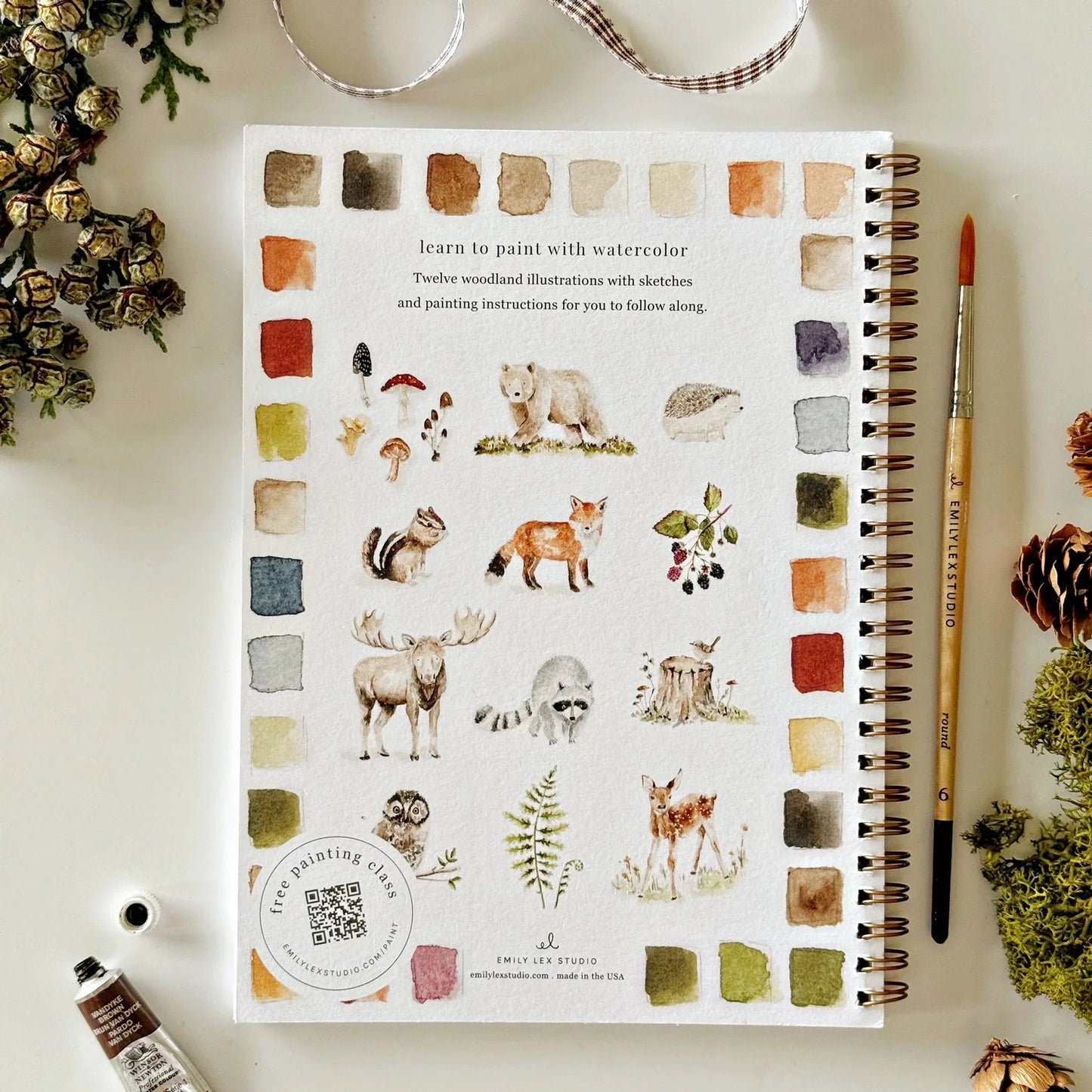 Notebook with watercolor woodland animal and plant illustrations and a paintbrush on a white surface