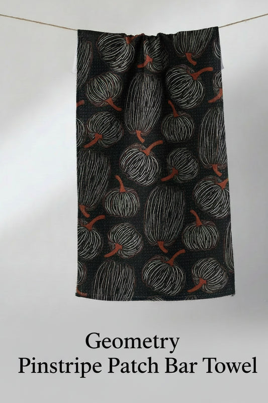 A black geometry pinstripe patch bar towel with a repeating pattern of white hand-drawn pumpkins and orange stems, hanging from a thin string against a light gray background.