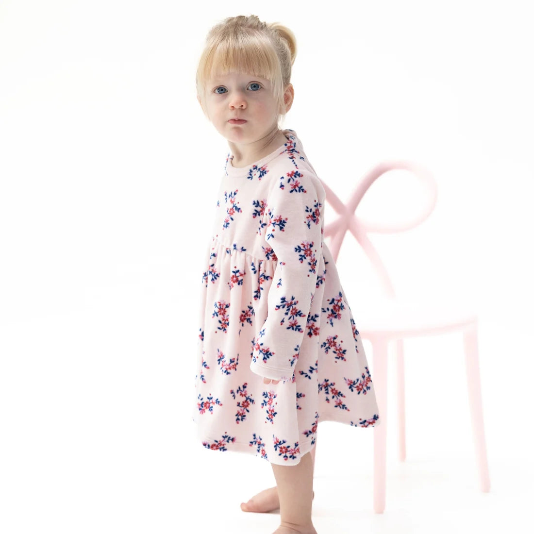 A toddler girl with blonde hair in a ponytail stands barefoot in front of a white background, wearing a light pink Angel Dear Nora Floral Velour long-sleeve tunic dress with a blue and red floral pattern. A light pink chair is visible behind her.