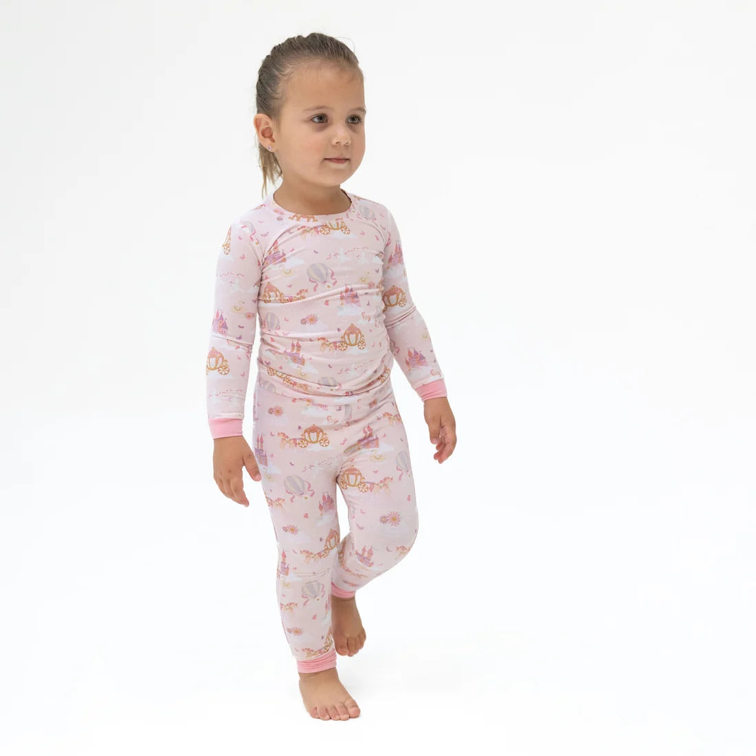 A young child wearing angel dear princess unicorn  light pink long-sleeve pajama set with a whimsical castle and fairy print, walking barefoot against a plain white background.