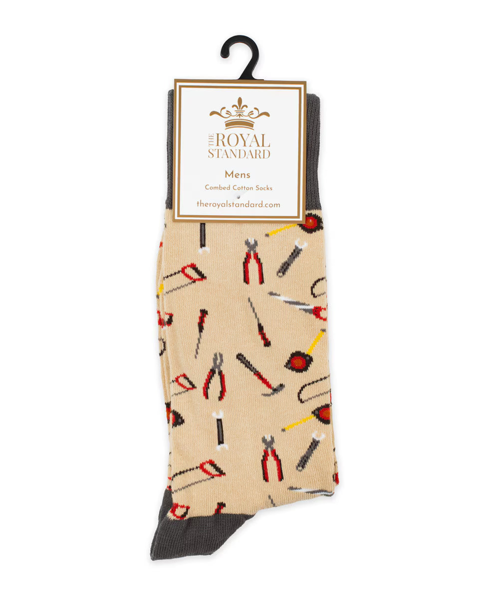 A pair of men's crew socks hanging from a black hanger with a paper label The Royal Standard. The socks are light beige with an allover pattern of hand tools including pliers, screwdrivers, and wrenches in red, black, white, and yellow.