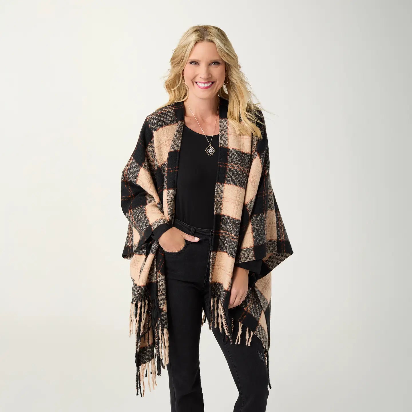 A woman modeling a black and beige plaid fringed wrap over a black outfit, standing against a light gray studio background.