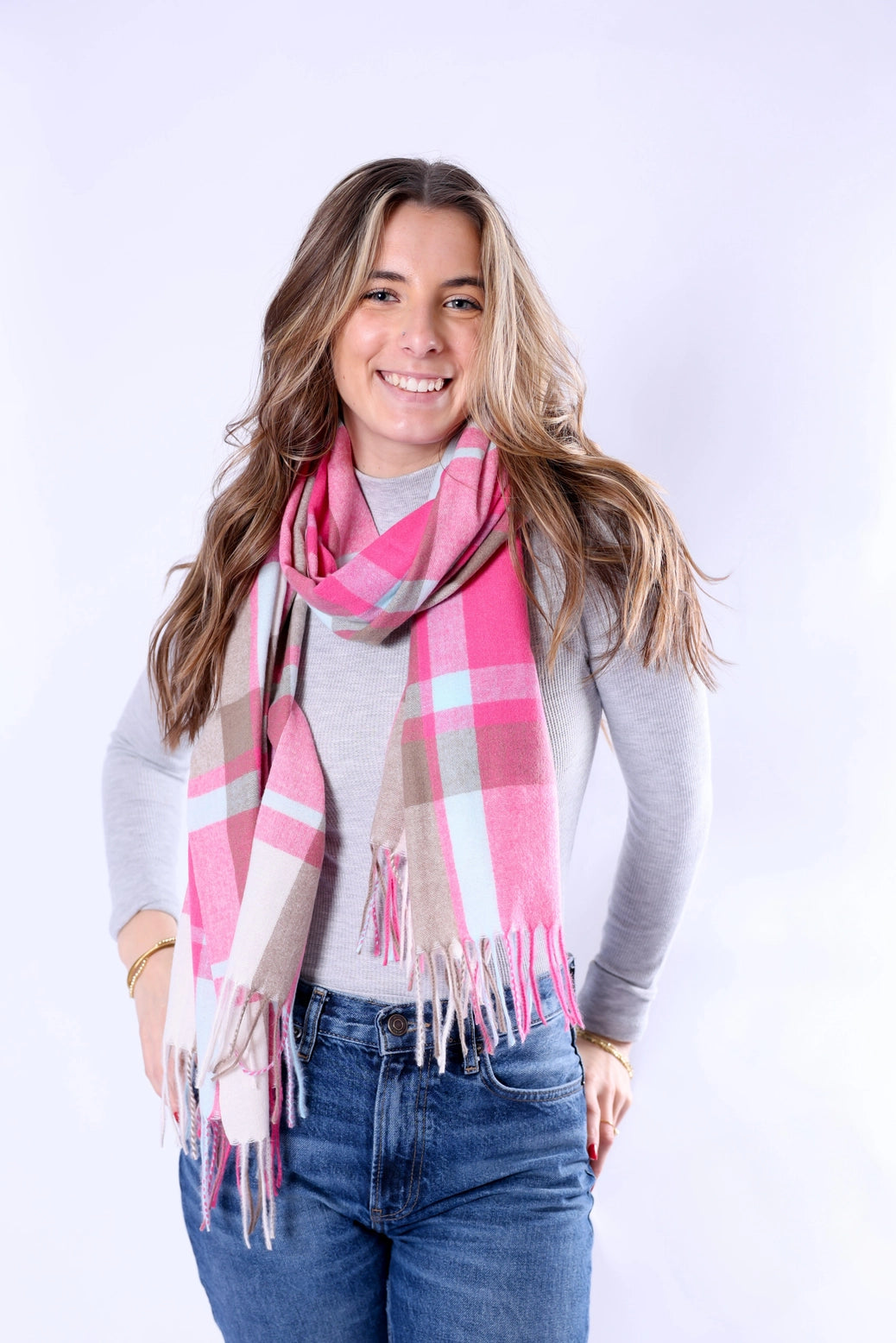 A smiling woman with long light brown hair models a pink, beige, and white plaid scarf with fringe details, paired with a gray long-sleeve top and blue jeans against a light gray studio background.
