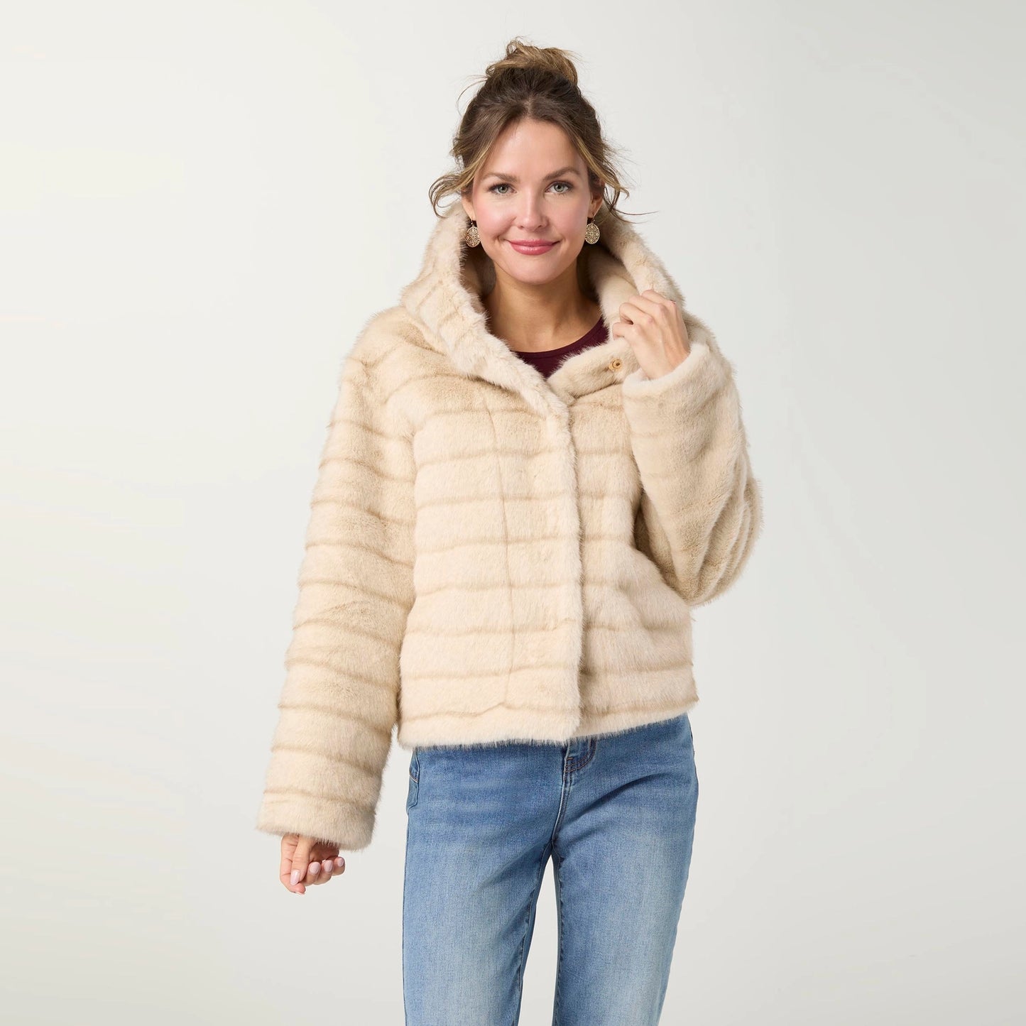 A woman modeling a cropped faux fur hooded jacket in a light beige color, paired with blue jeans, against a plain light gray studio background.