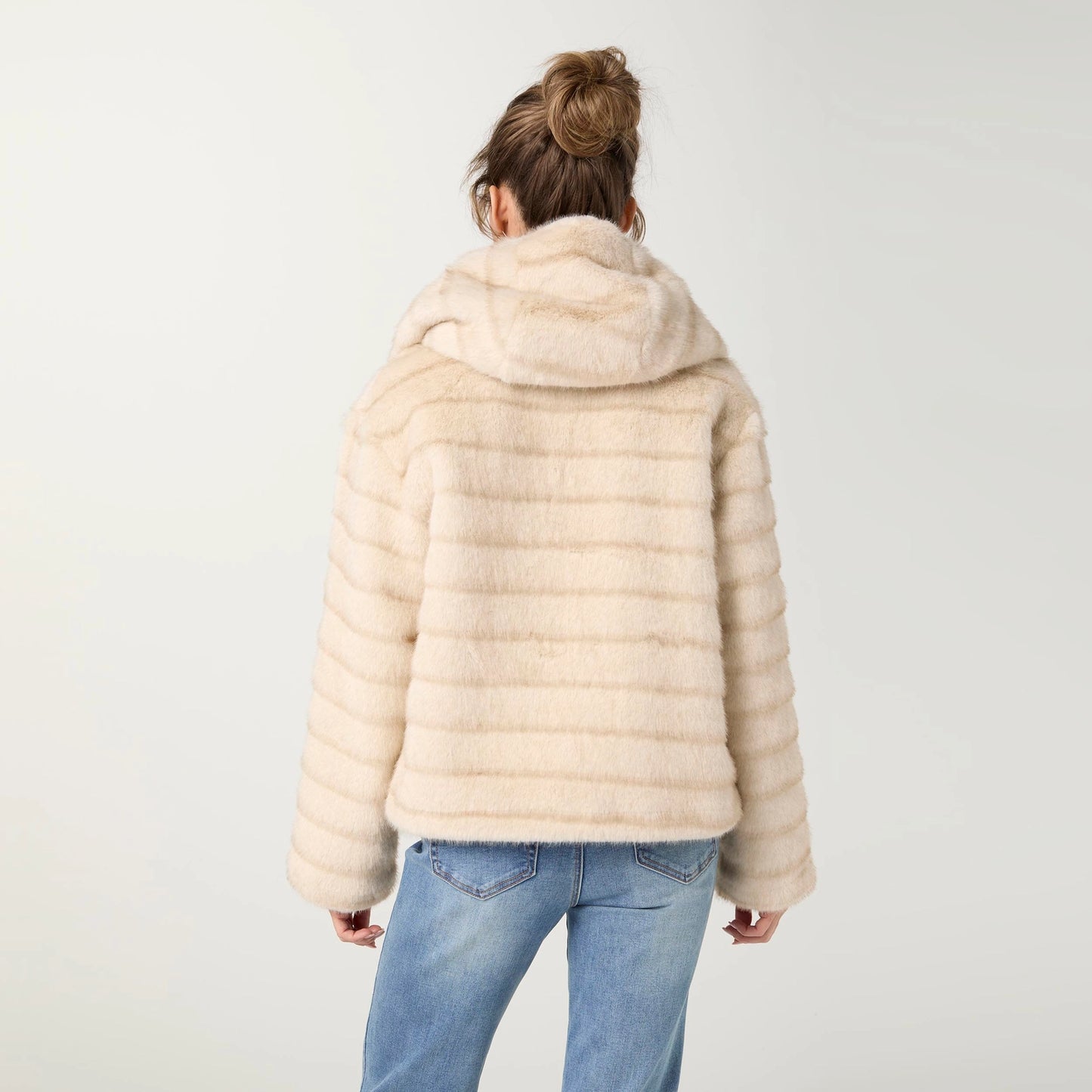 A woman viewed from behind wearing a light beige faux fur hooded jacket with horizontal ribbed sleeves, paired with blue jeans, against a plain light gray background.